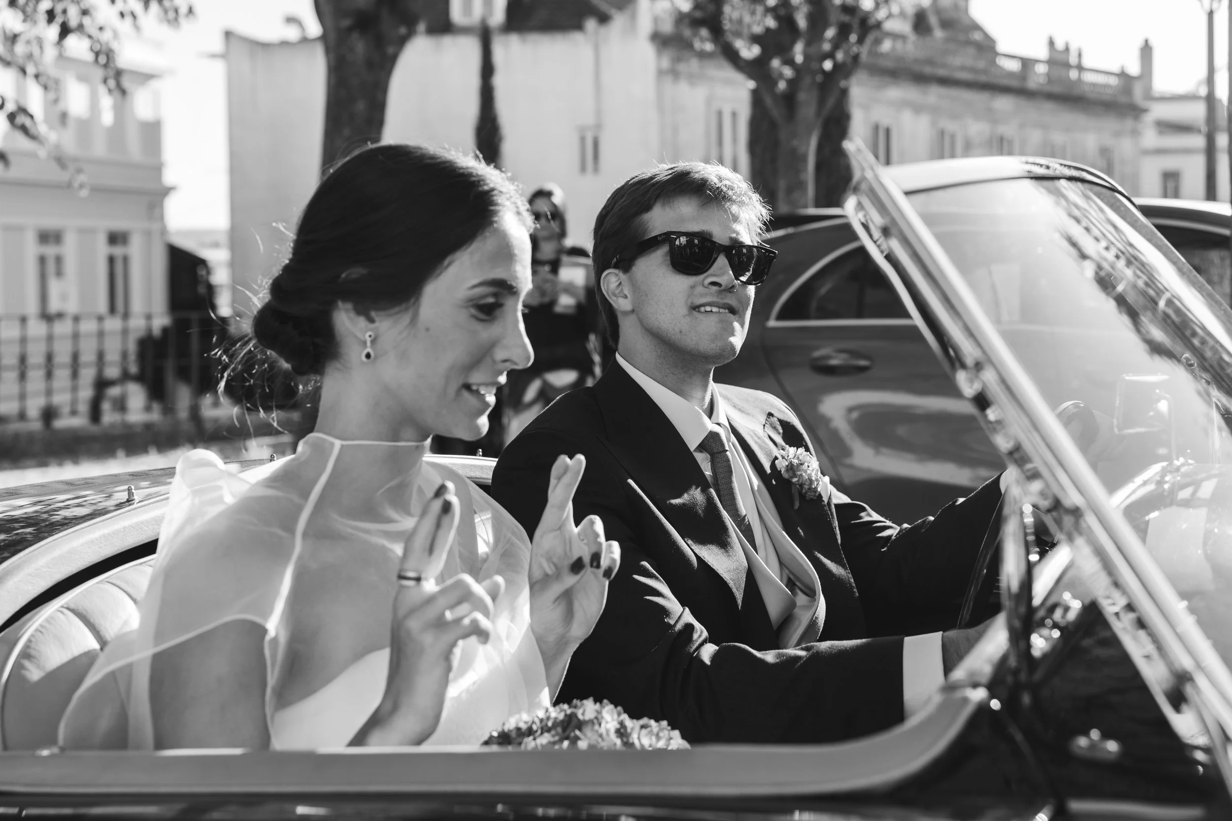 A black and white photo of a bride and groom driving in a vintage convertible car. The bride has dark hair styled elegantly, and the groom is wearing sunglasses and a suit. They seem happy, possibly on their wedding day.