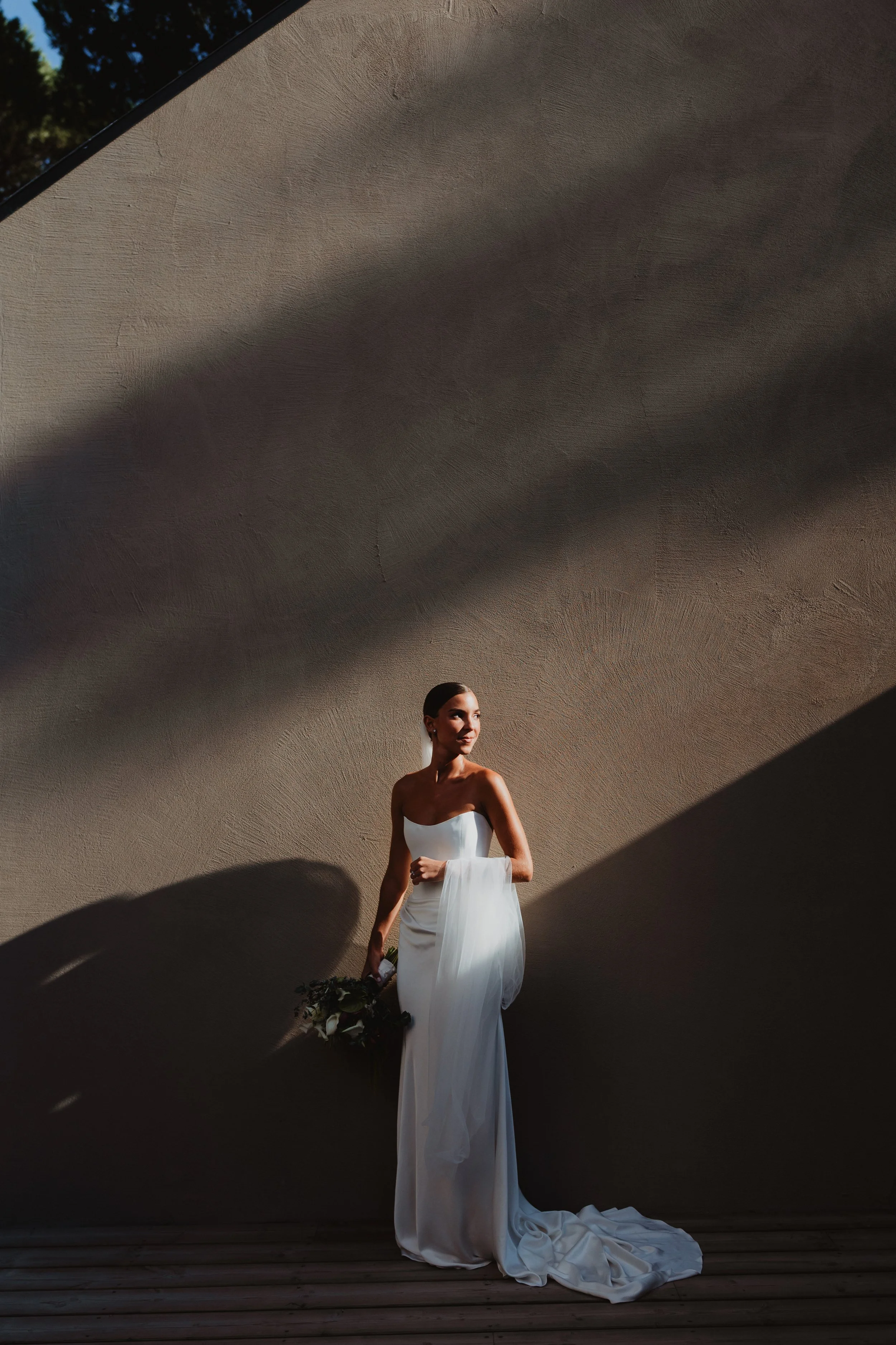 A woman in a white strapless wedding gown holding a bouquet, standing outdoors in front of a textured gray wall with dramatic shadows.