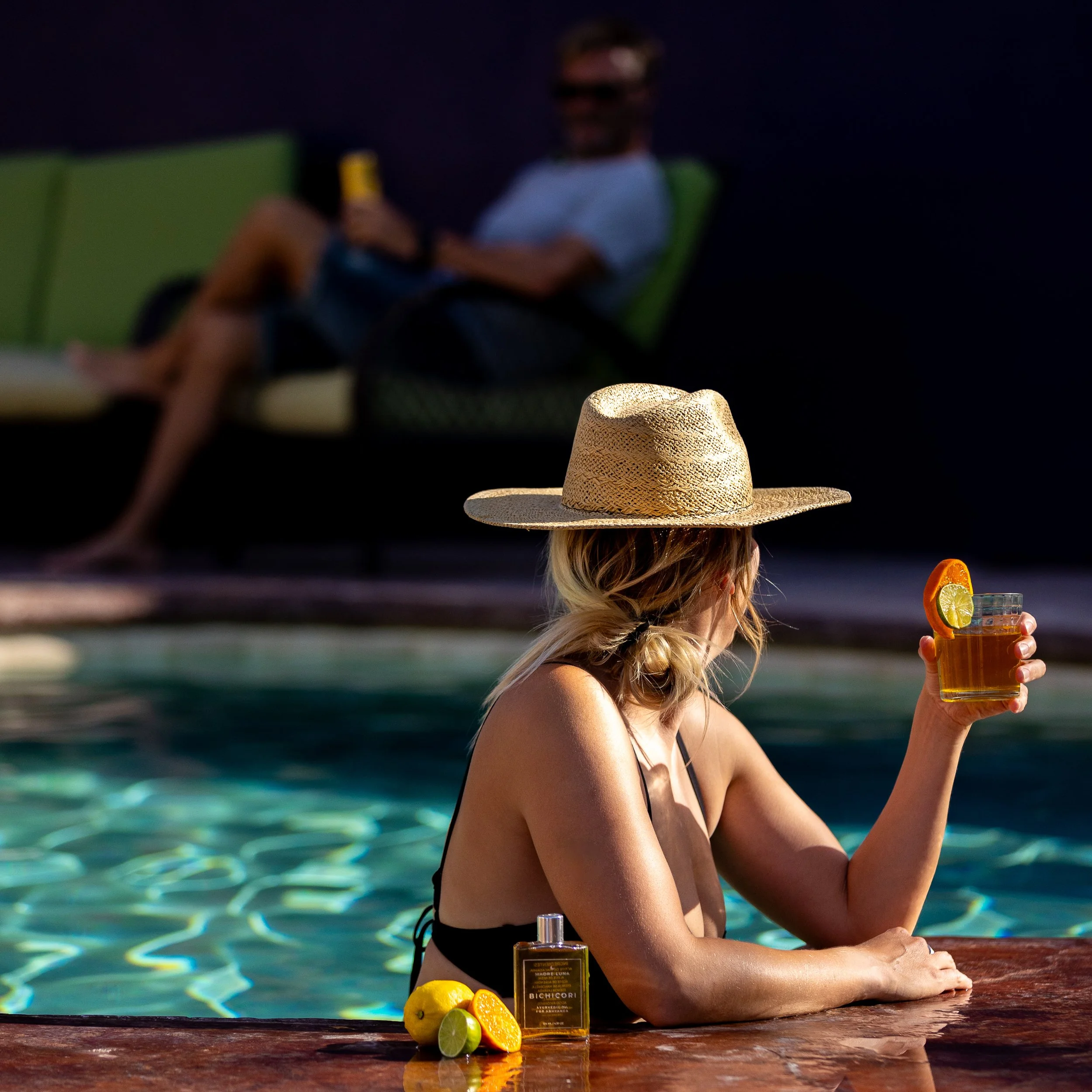 A hotel guest wearing a wide-brim hat relaxing by a pool and holding a cocktail. There is another guest lounging in the background at a boutique hotel in Los Barriles, Baja California Sur, Mexico.