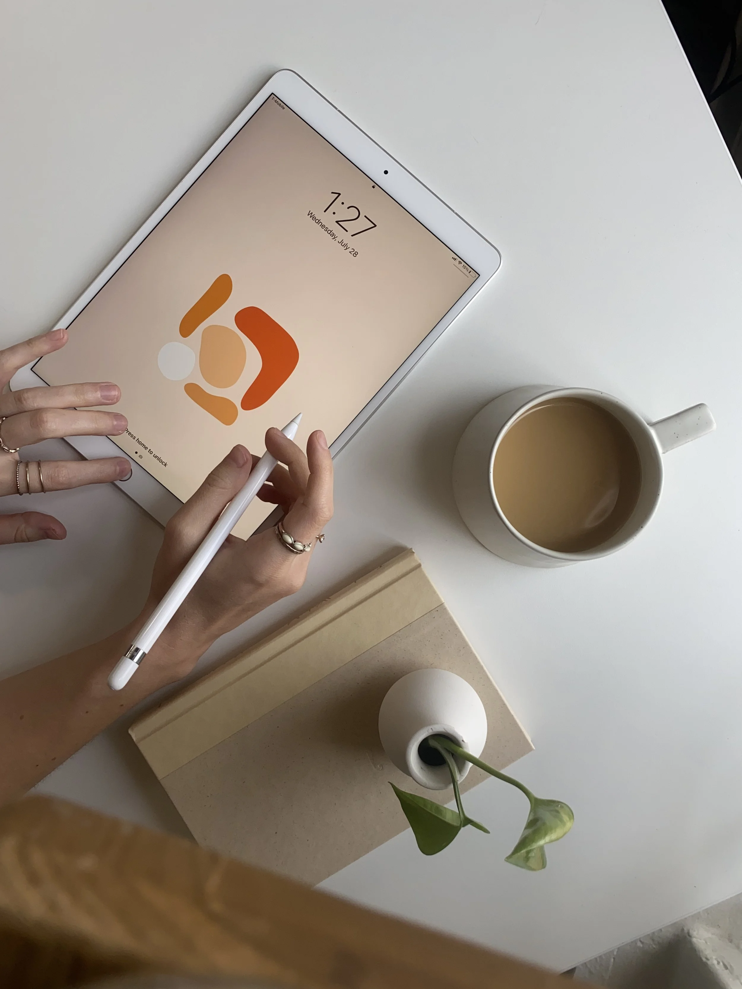 An overhead view of a person using a white tablet with an Apple Pencil, a beige notebook, a white mug filled with coffee, and a small white vase with a single green flower on a white table.