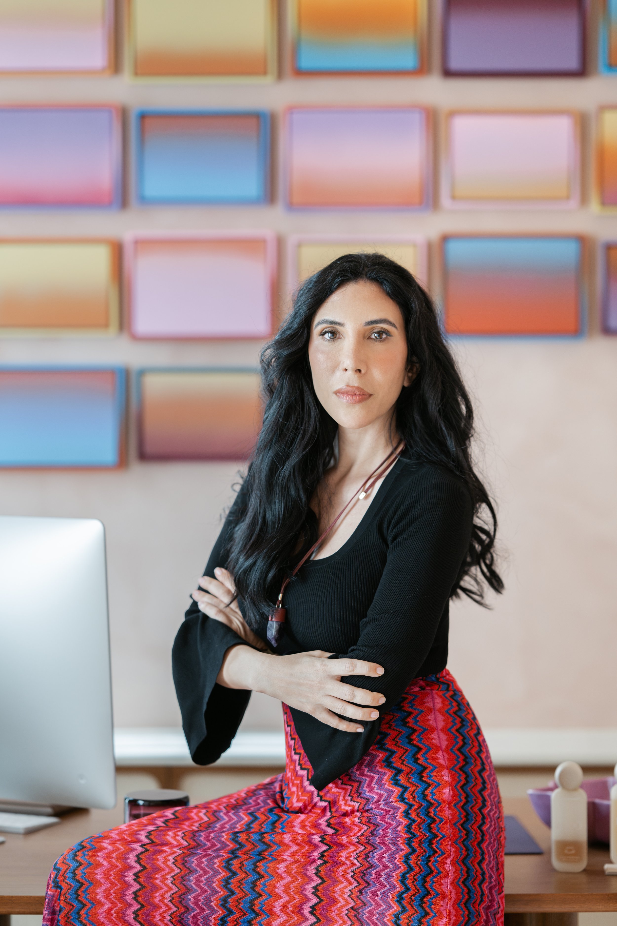 A woman with long dark hair, wearing a black top and a colorful red and pink patterned skirt, sitting on a desk with her arms crossed, in front of a wall with pastel-colored artwork. Various items including a computer, a mug, and a bottle are on the desk.