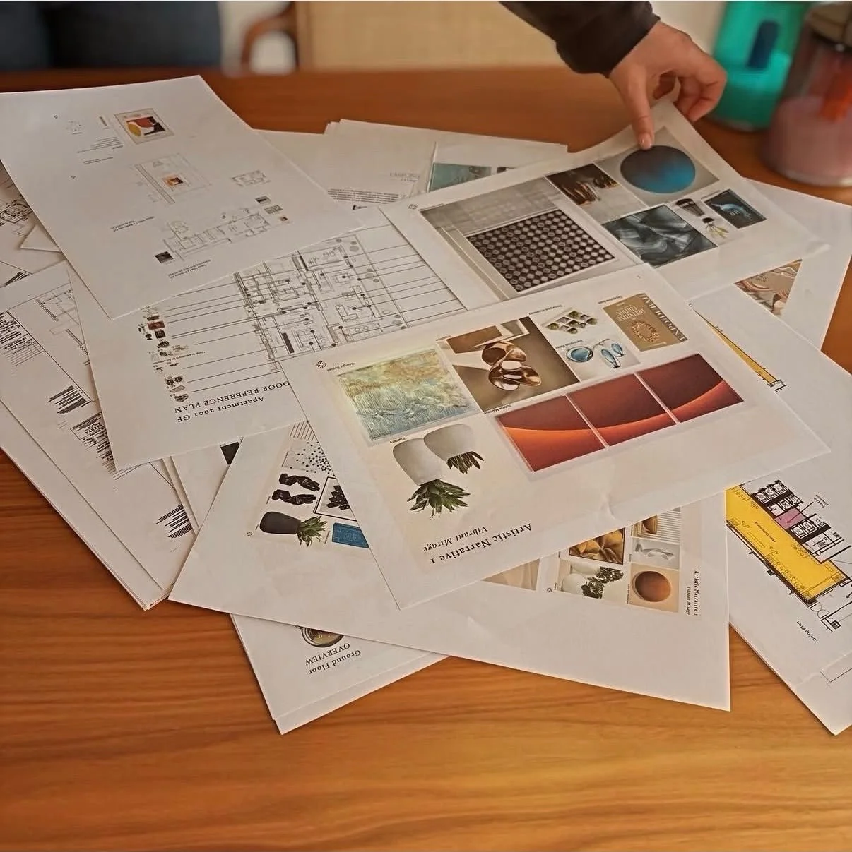 A person sorting through various papers and design plans on a wooden table, including interior design layouts, magazine pages, and architectural drawings.