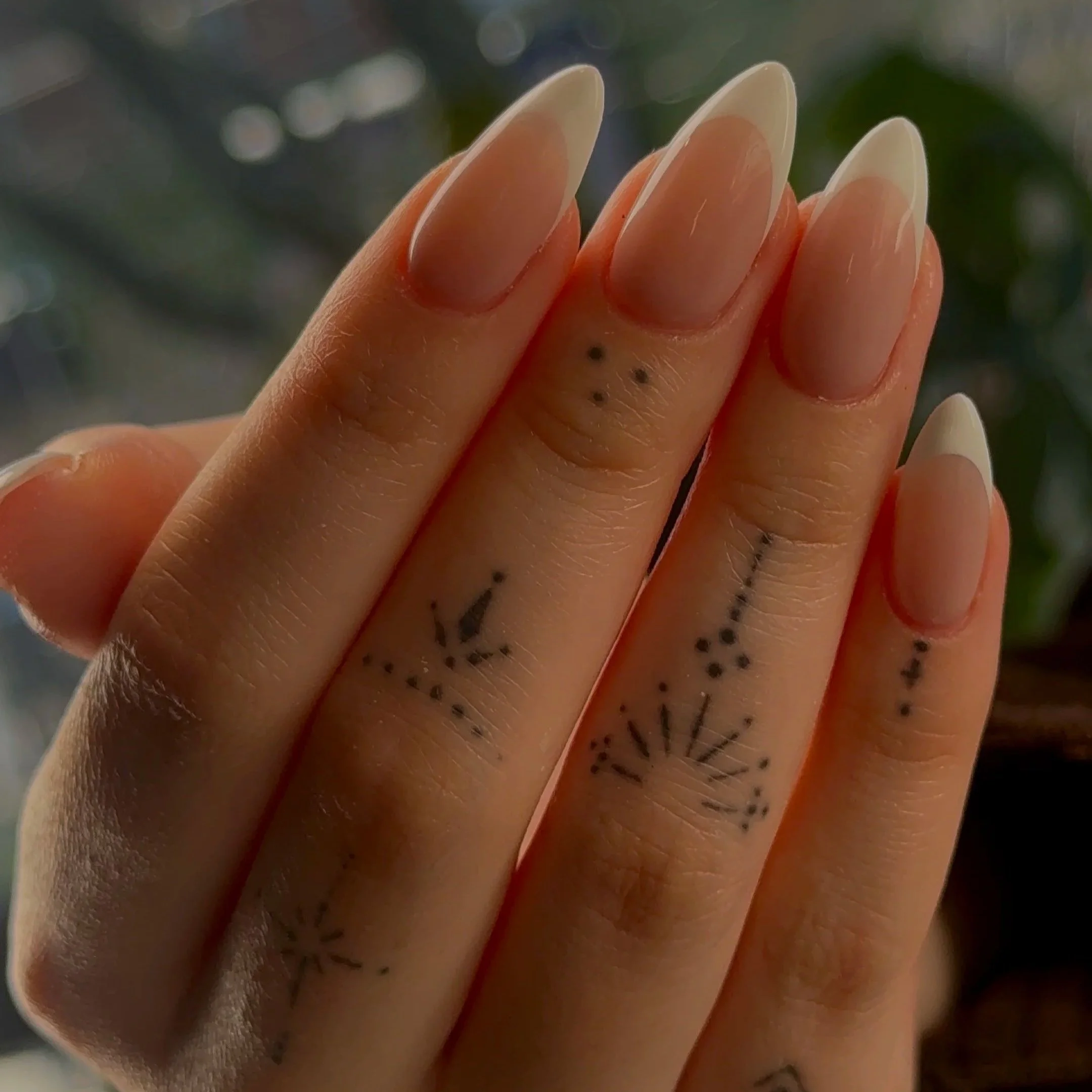 A close-up of a person's hand with natural nails and small black freckles or dots, with a blurred background of green and gray tones.