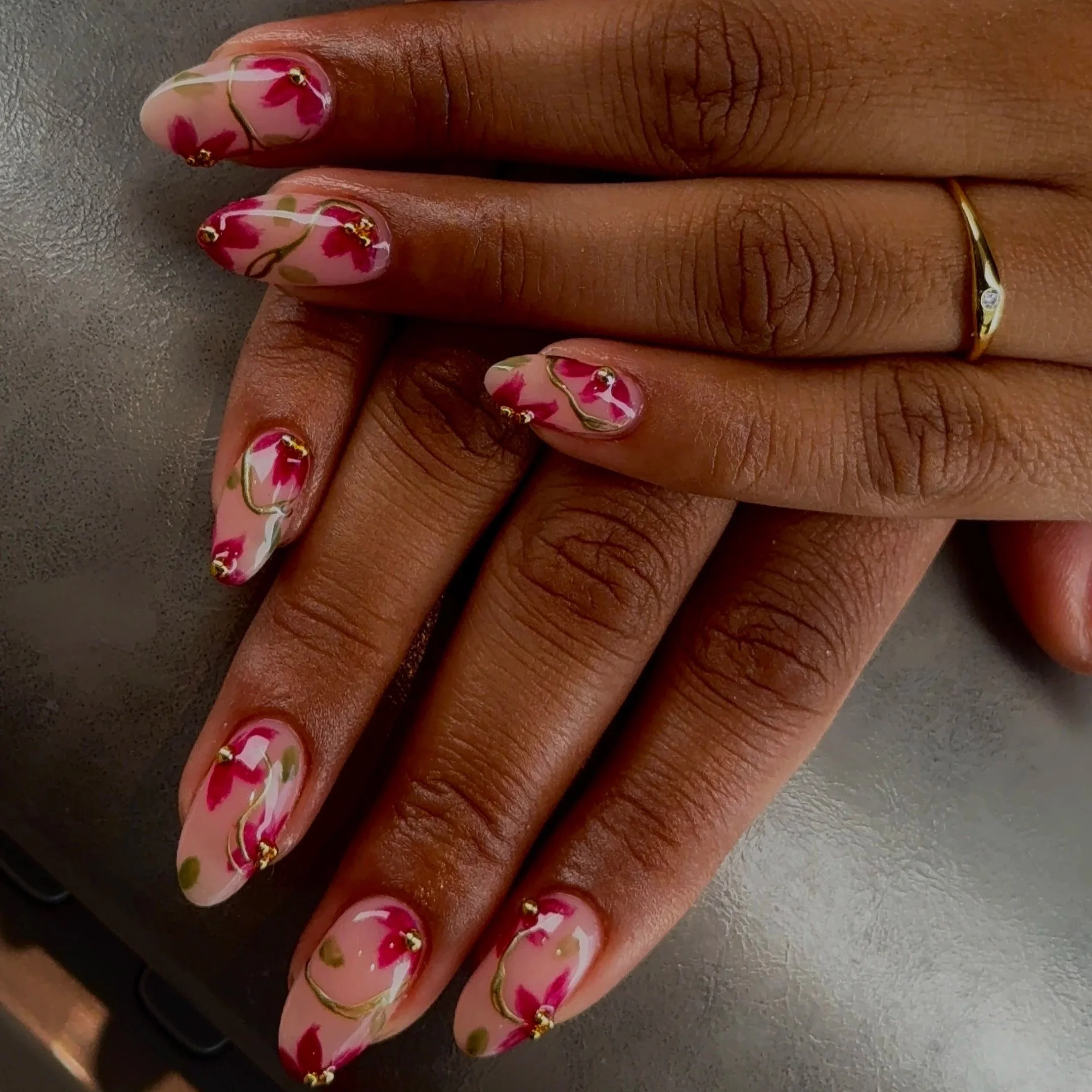Close-up of a woman's hand with nails painted in floral design, featuring pink, white, and green colors with gold accents, and wearing a gold ring with a small gemstone.