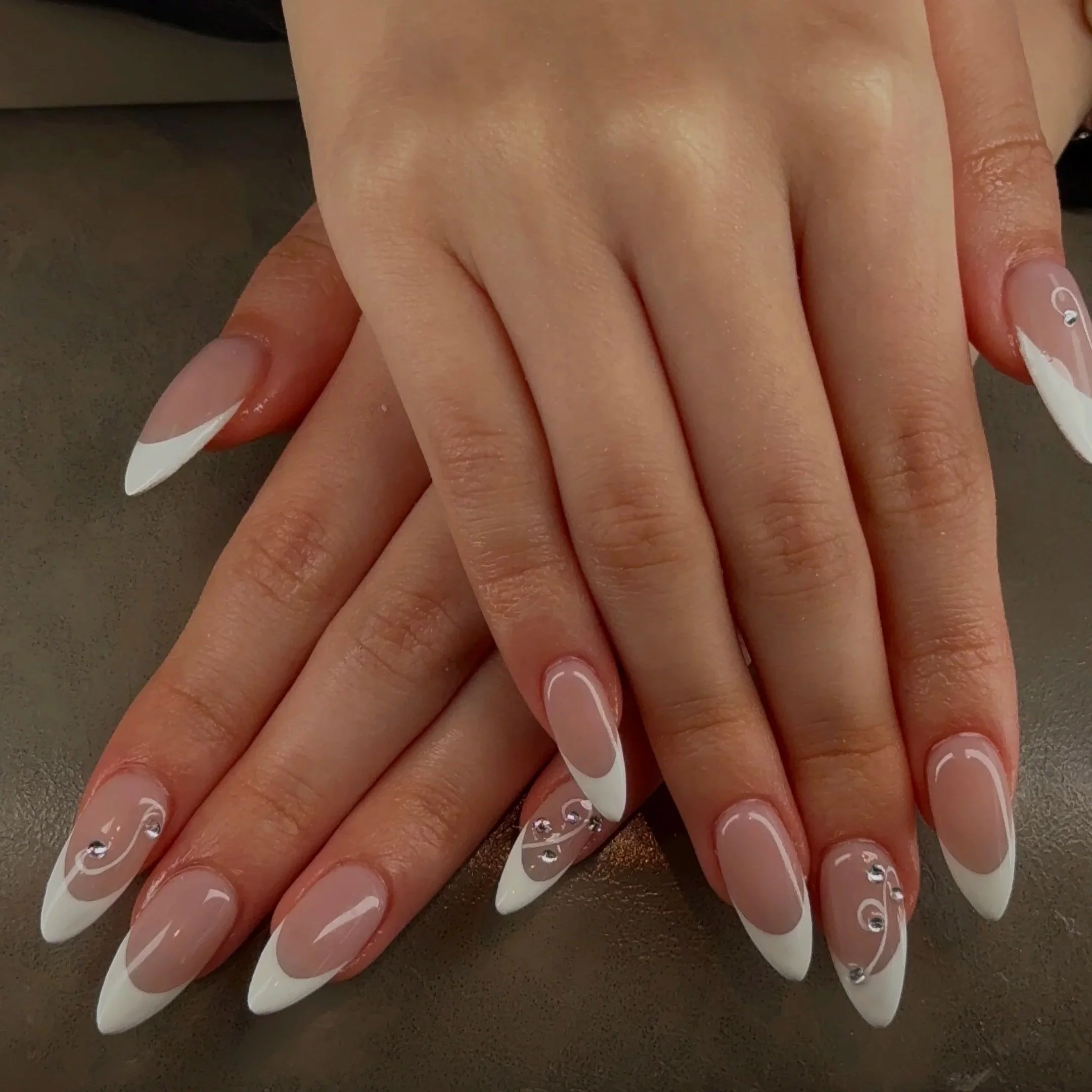 Close-up of manicured fingernails with white French tips and small rhinestones on a person's hand, resting on a dark surface.