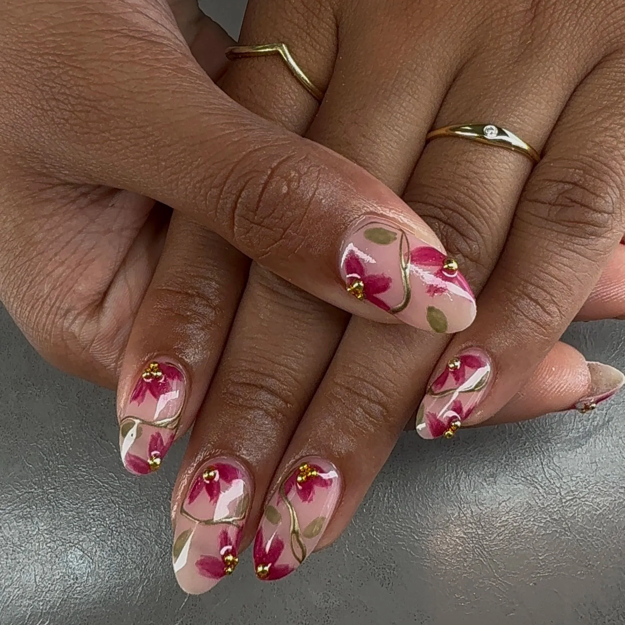 Close-up of a person's hand with gold and diamond rings, featuring pink nail polish with floral designs and gold dots, resting on a textured surface.
