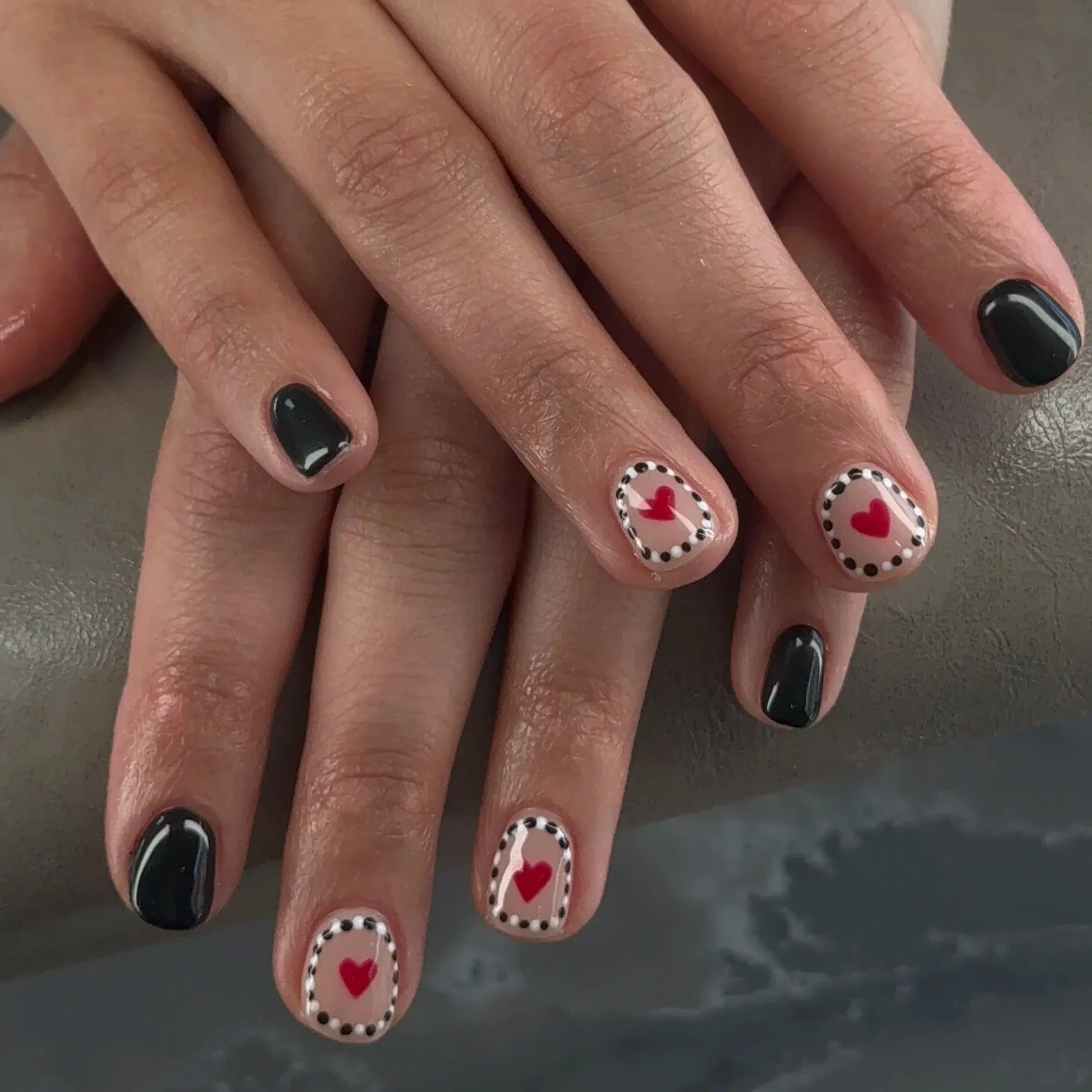 Close-up of a person's hand with black nail polish, featuring decorated nails with red hearts, white borders, and black dotted borders on a pale background.