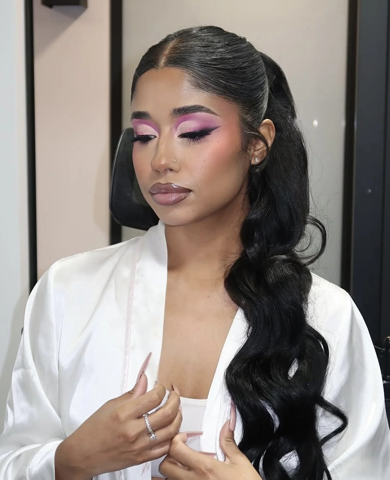A woman with long black wavy hair, makeup with pink eyeshadow and winged eyeliner, and nude lipstick, looking down with her eyes closed. She's wearing a white satin robe and holding her chest with both hands, showing a ring on her left ring finger.