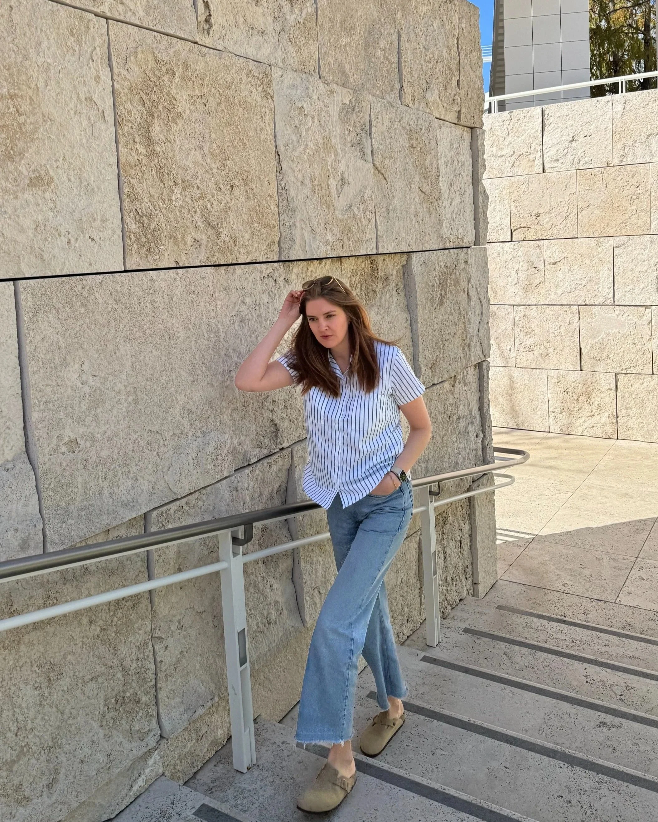 A woman stands on a stone walkway next to a textured beige wall, wearing a striped blouse, blue jeans, and beige shoes. She is leaning against the railing and touching her hair, with modern architectural elements in the background.