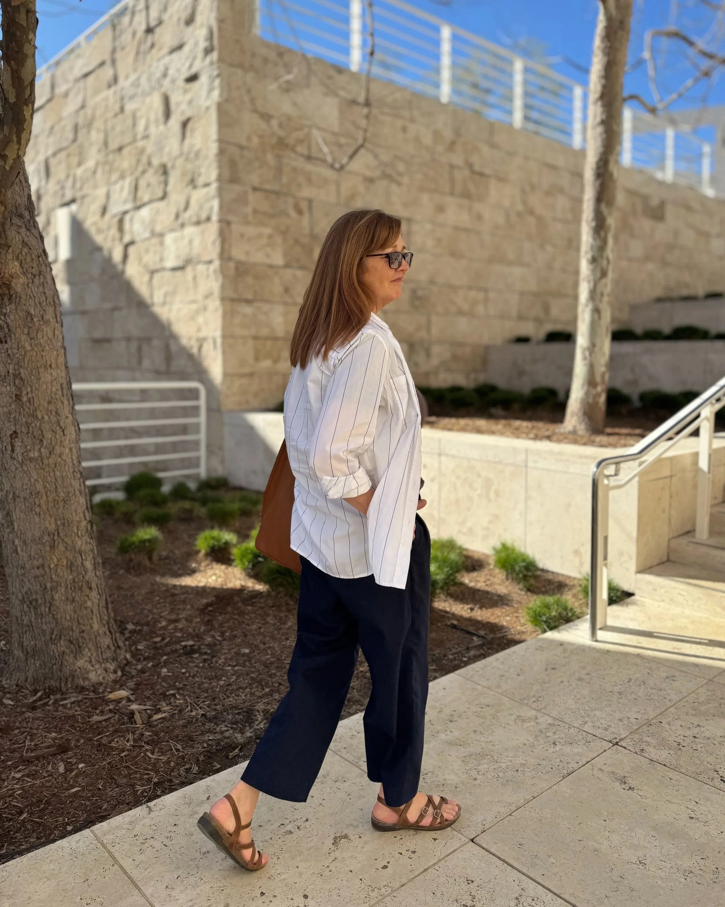 Woman walking outdoors wearing a striped shirt, dark pants, sandals, and sunglasses, with a brown bag. Modern architectural background and clear sky.