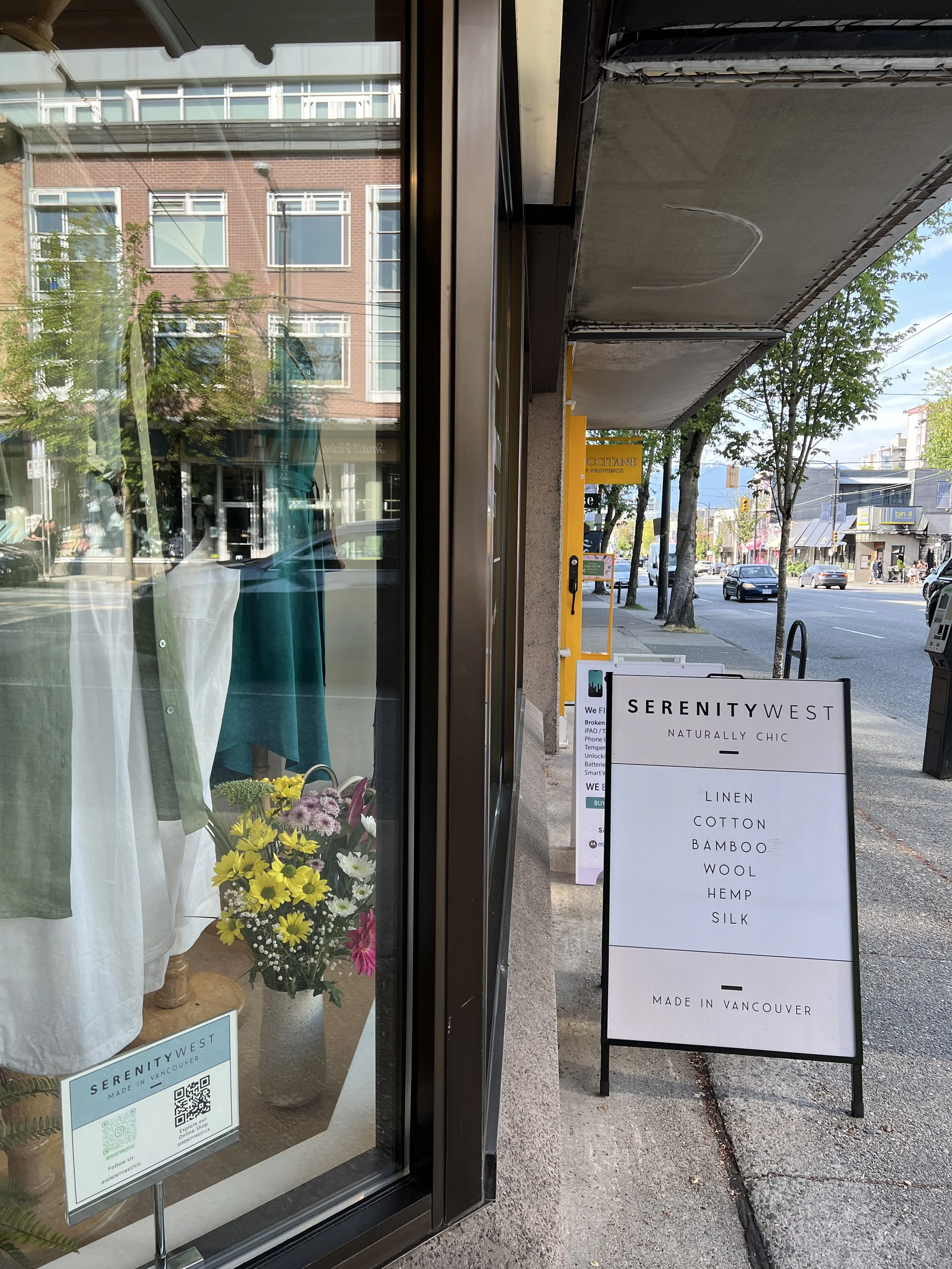 Storefront of Serenity West in Vancouver with a sidewalk sign listing materials like linen, cotton, bamboo, wool, hemp, and silk. The display window shows clothing and a bouquet of flowers.