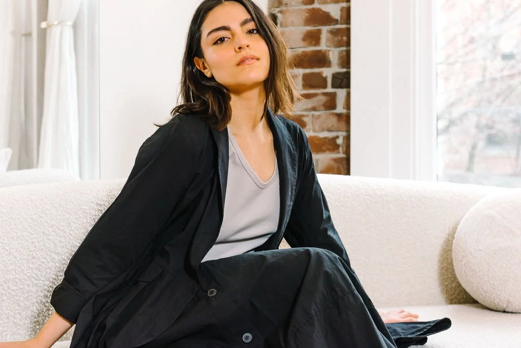 Woman sitting on a light-colored couch wearing a black outfit, with a brick wall and window in the background.