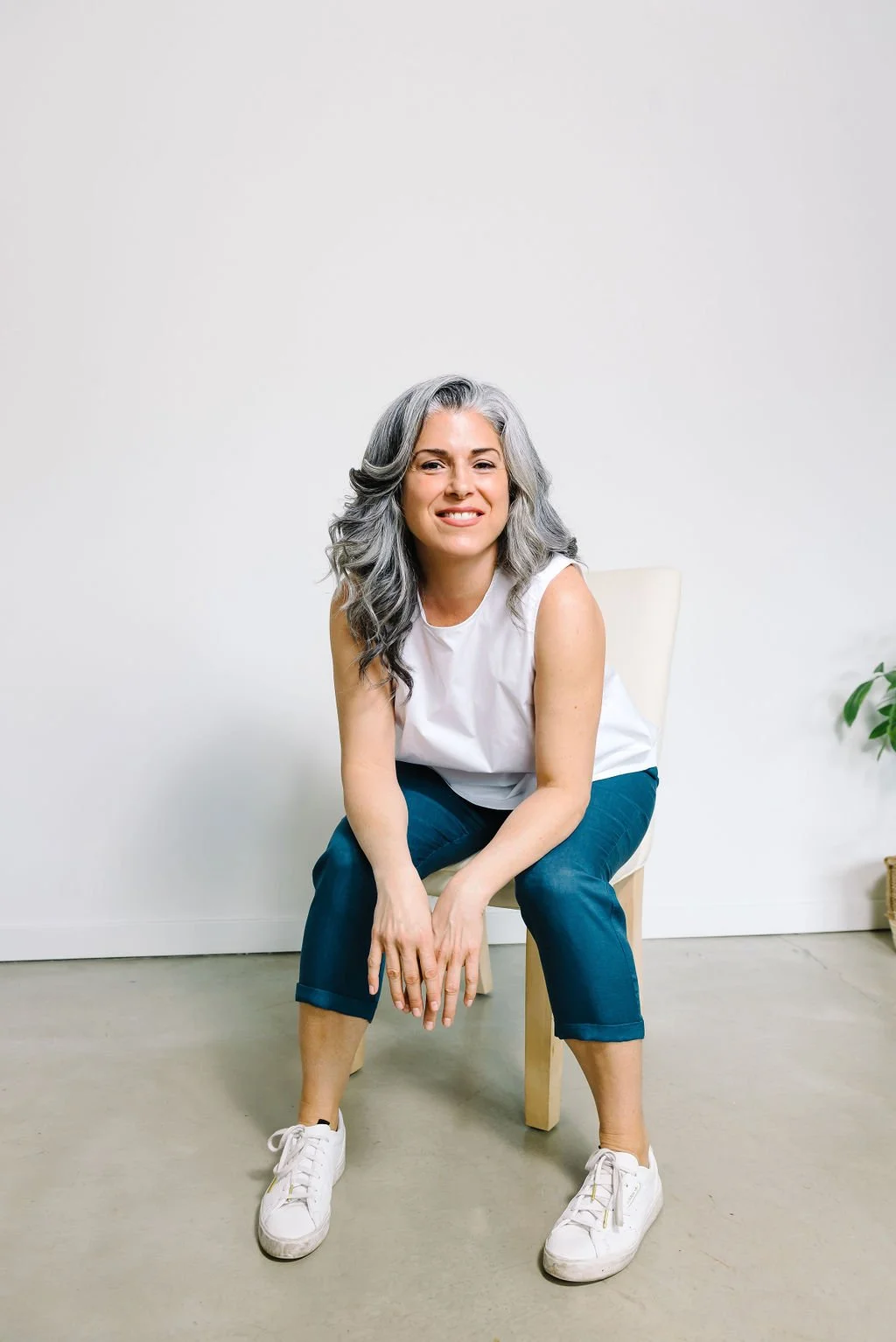 Woman with gray hair sitting on a chair, wearing a white sleeveless top, blue pants, and white sneakers, smiling in a minimalistic room.