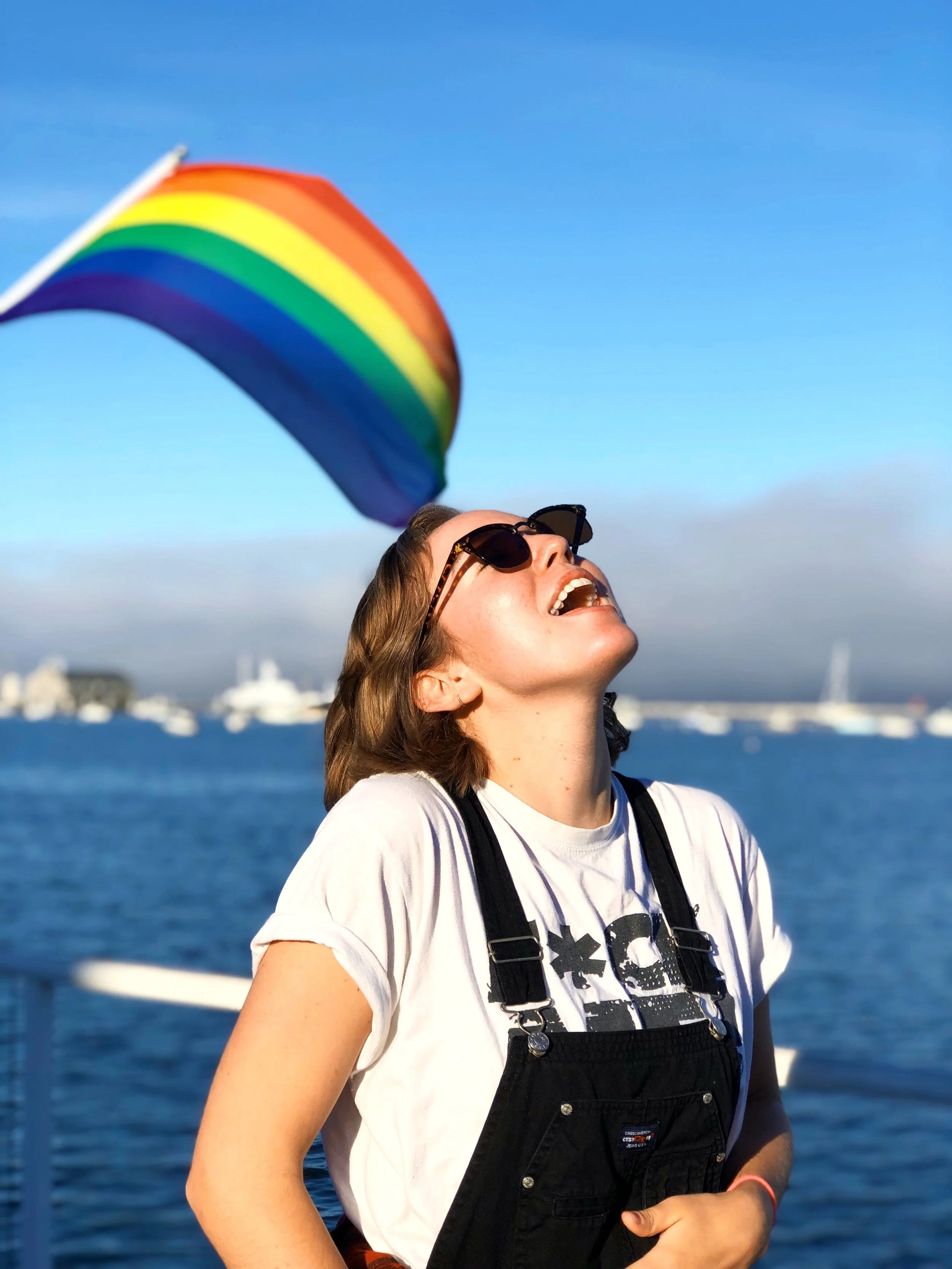 Young woman in sunglasses smiling and laughing outdoors near water with a rainbow flag and boats in the background.
