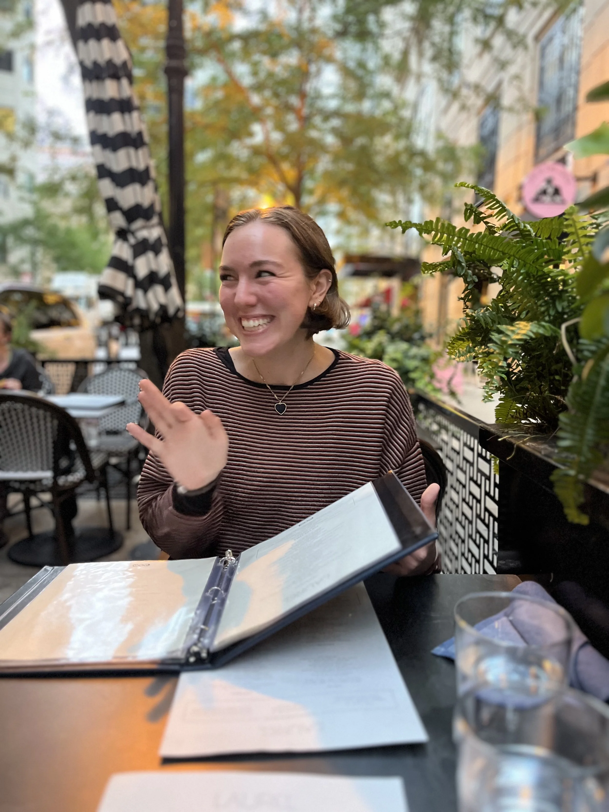 A woman with short brown hair smiling and laughing, sitting at an outdoor cafe table, holding a menu in front of her, with glasses of water and papers on the table, surrounded by greenery and outdoor city scenery.