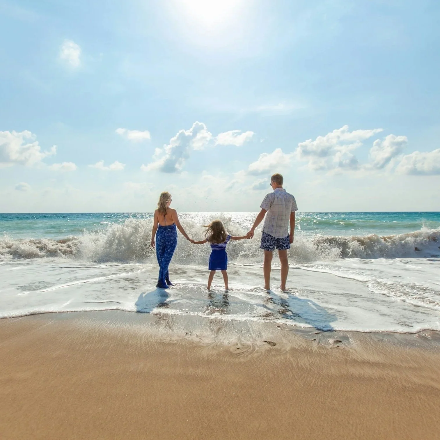 A family of three holding hands in the ocean waves on a sunny beach, with a blue sky and scattered clouds.