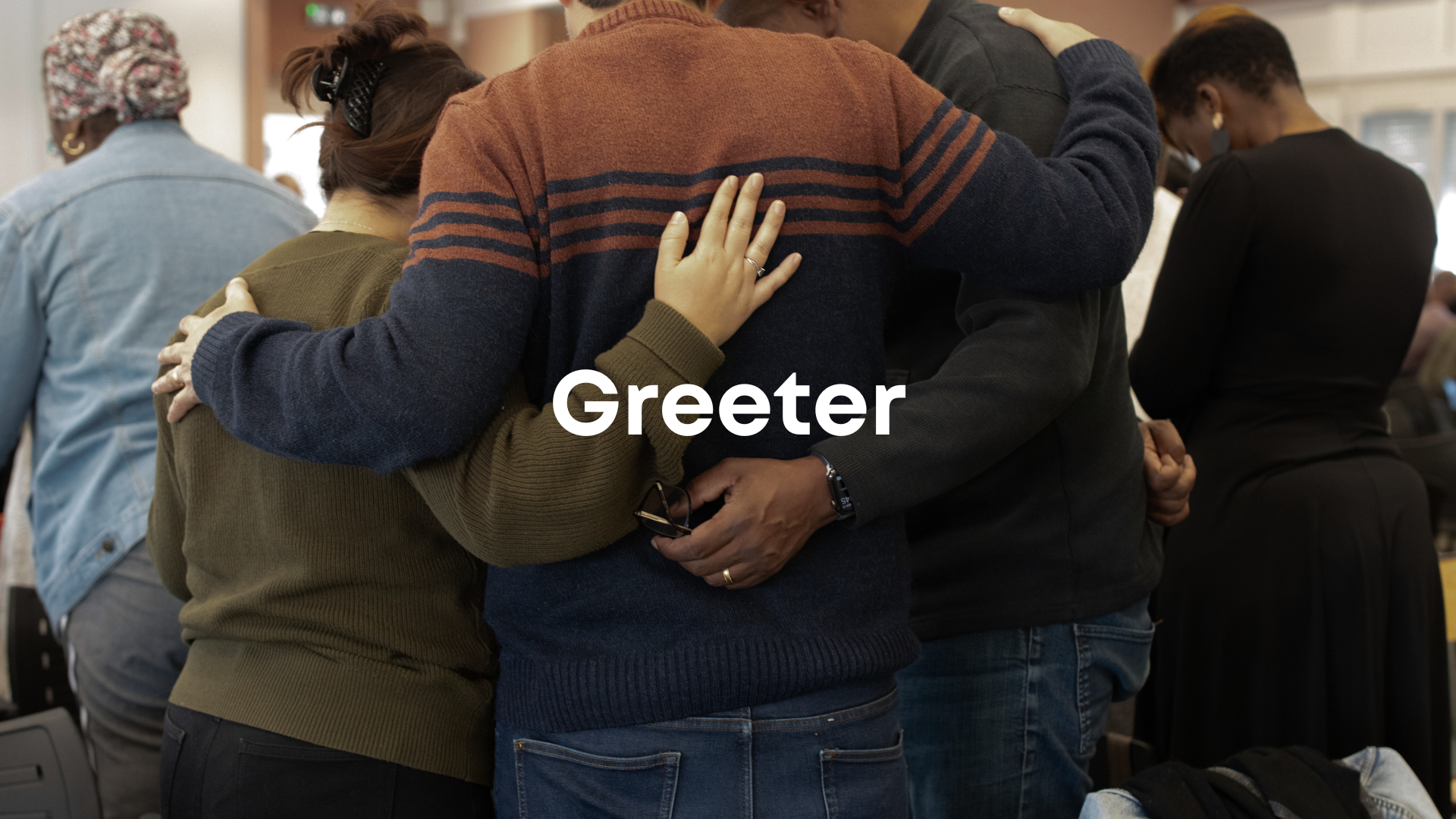 A diverse group of people embracing in a prayer circle, with the word 'Greeter' overlayed.