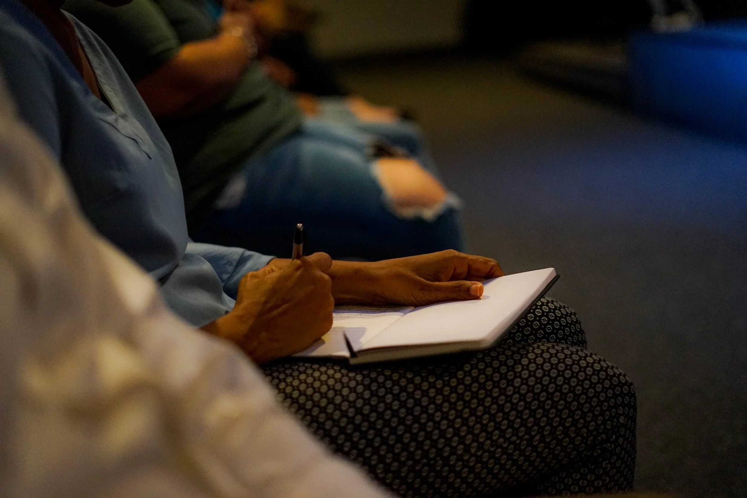 Close-up of a person sitting on a dark theater seat taking notes in a notebook with a pen.