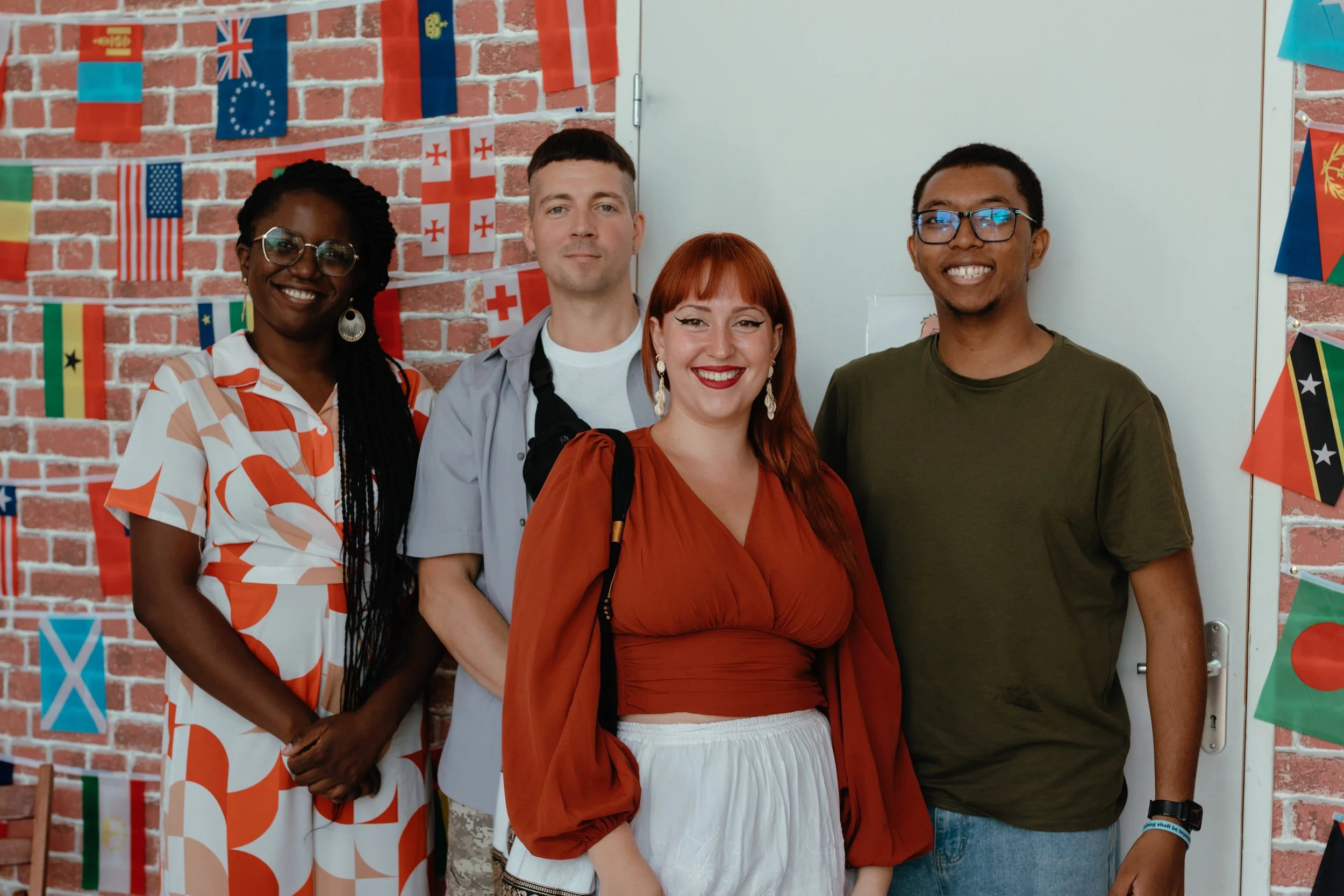 A diverse group of four young adults standing together in front of a brick wall decorated with international flags, smiling at the camera.