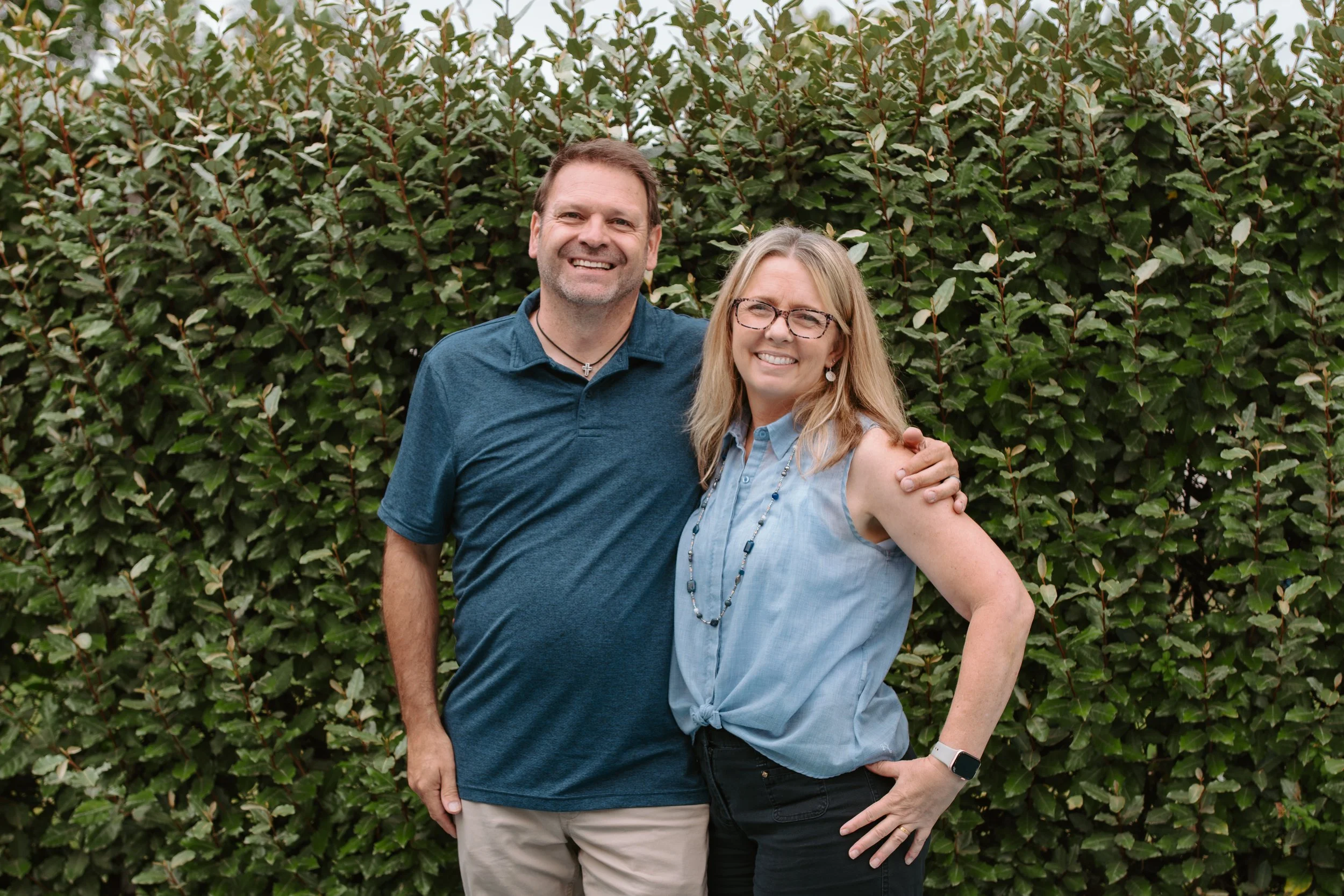 A smiling man and woman standing outdoors in front of a green hedge. The man has short brown hair, beard, and is wearing a blue polo shirt and a cross necklace. The woman has blonde hair, glasses, and is wearing a light blue sleeveless vest and a beaded necklace.