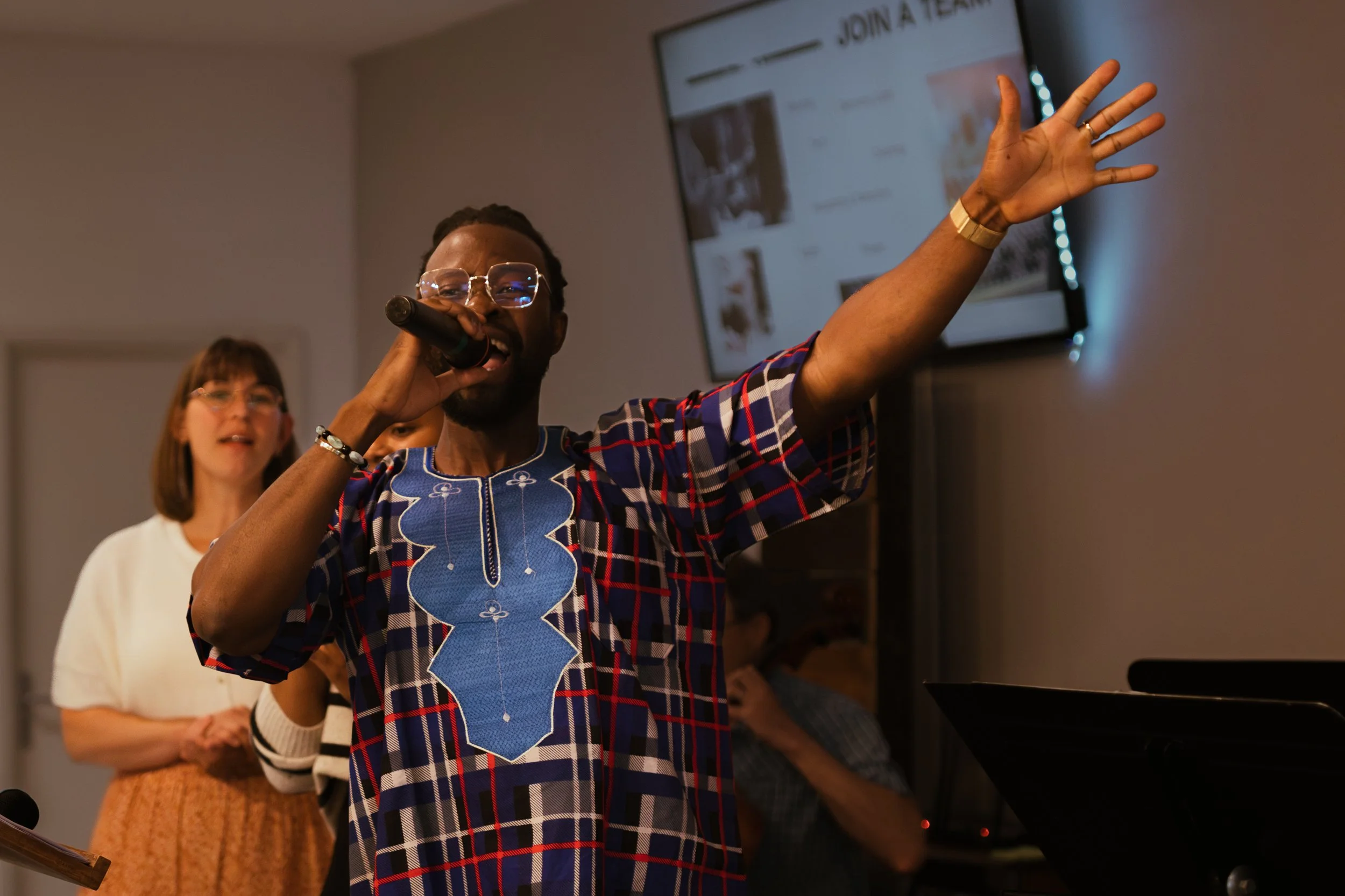 A man singing into a microphone, wearing glasses and a colorful patterned shirt, raising his hand, with two women in the background, one wearing a white top and glasses.