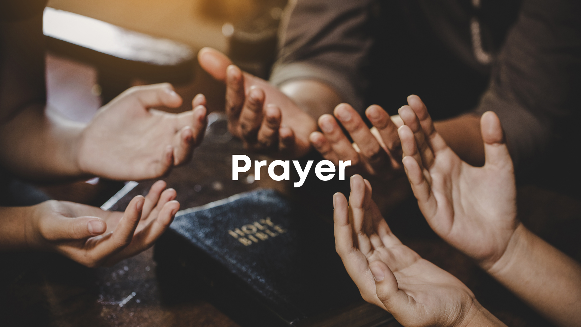 Multiple hands in various positions of prayer over a Bible, with a warm soft focus background.