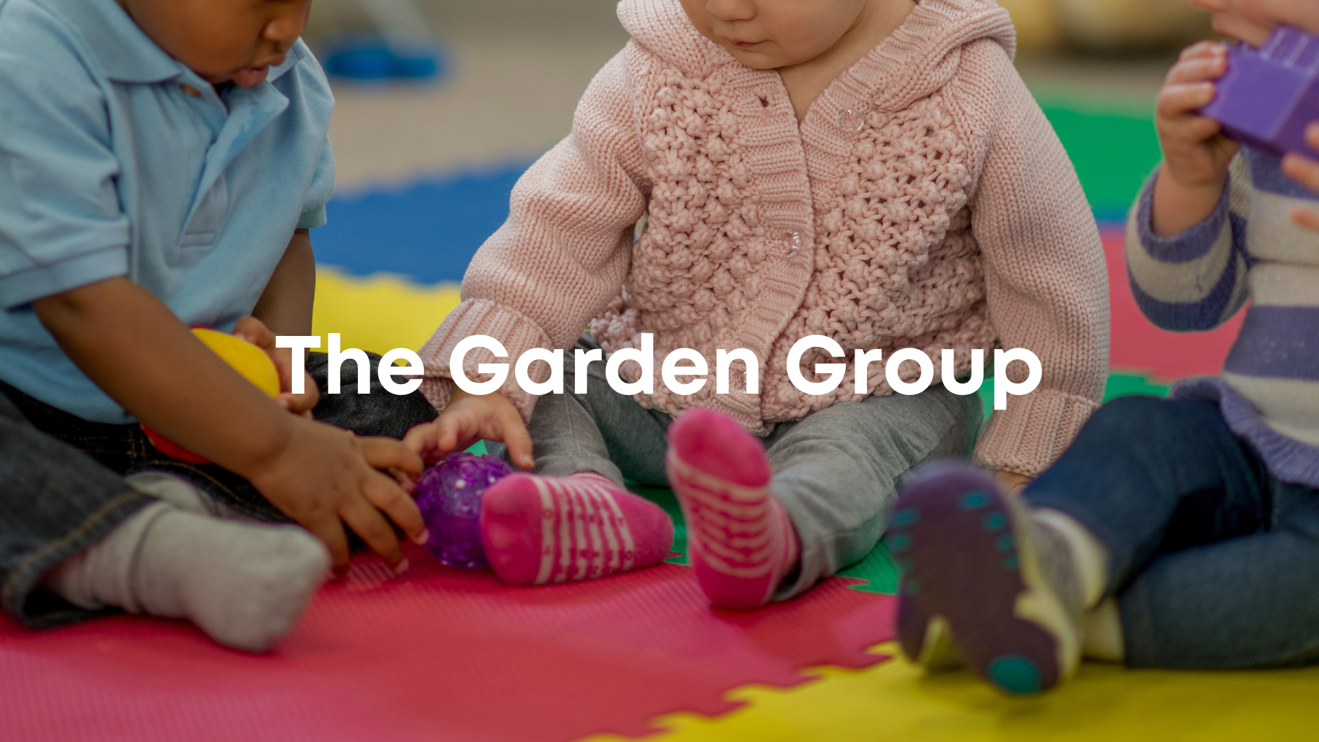 Close-up of young children sitting on colorful foam mats, engaging in a group activity, with the text "The Garden Group" overlaid.