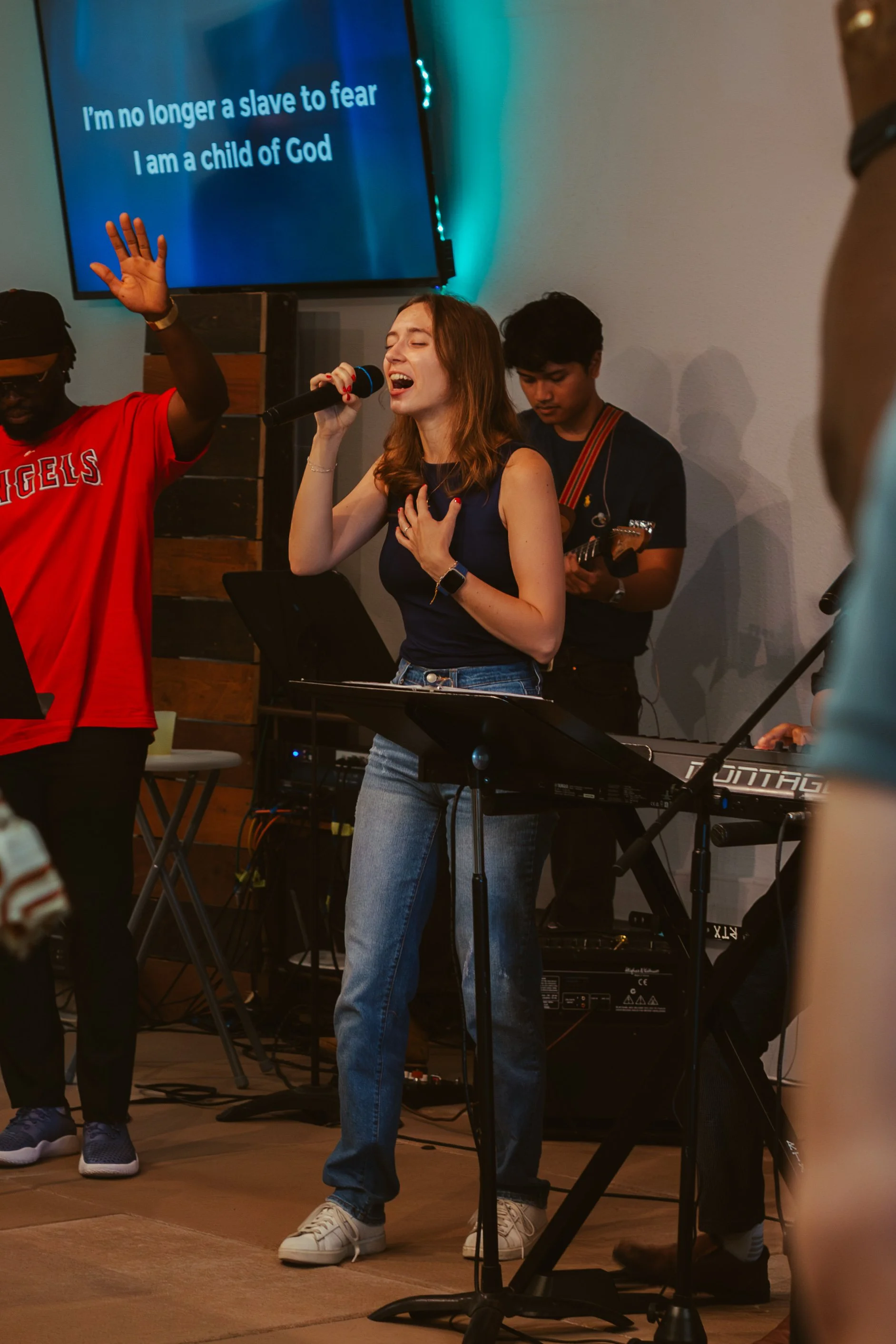 A woman singing into a microphone with her hand on her chest, surrounded by musicians and a large screen behind her displaying the lyrics, 'I'm no longer a slave to fear, I am a child of God.'