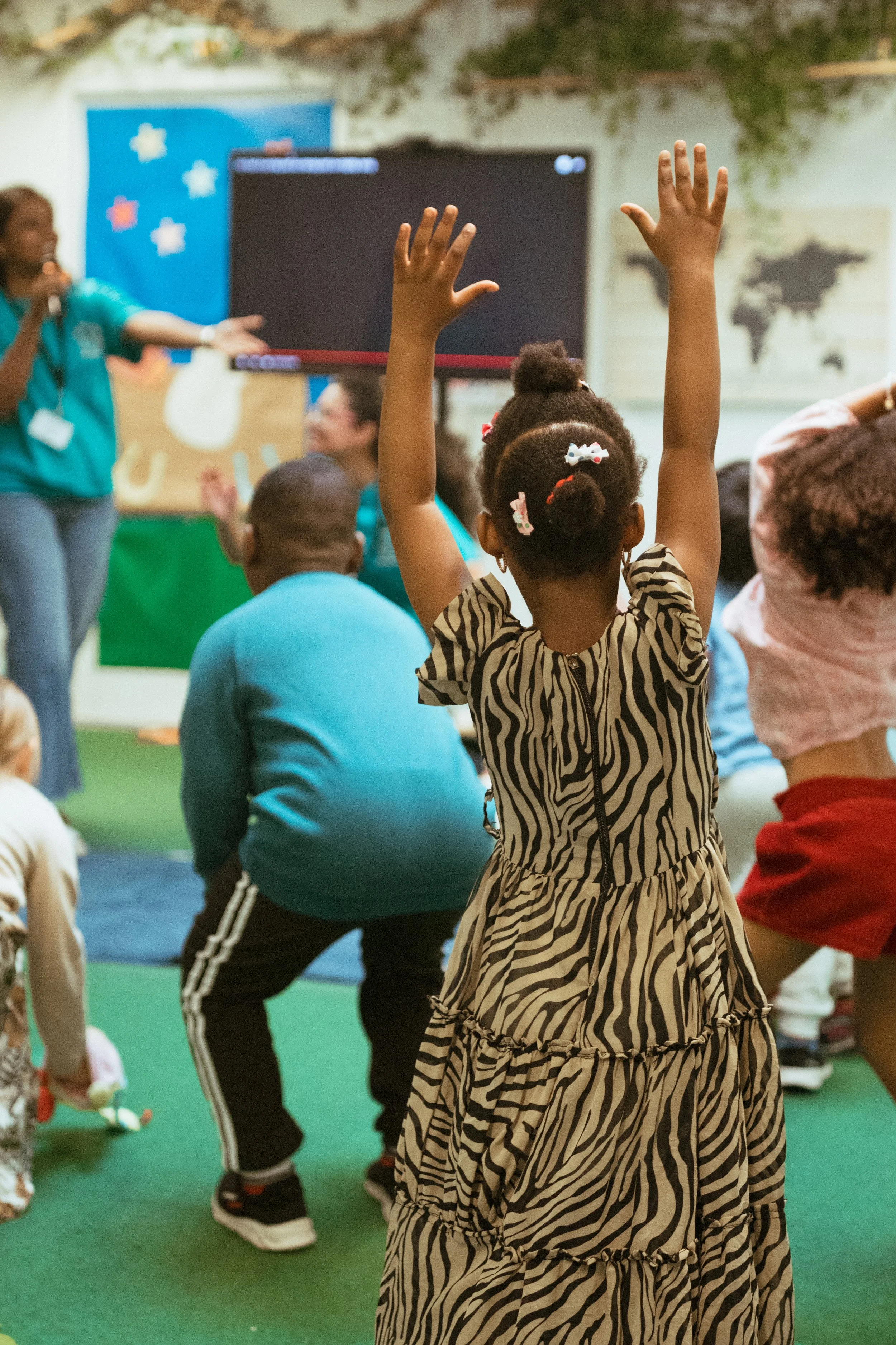 A young girl with curly hair tied up with white, pink, and red hair ties raising her hands during a classroom activity. She is wearing a zebra print dress. Other children are sitting on the floor, and a teacher is speaking in the background, with a large screen and a wall map visible.