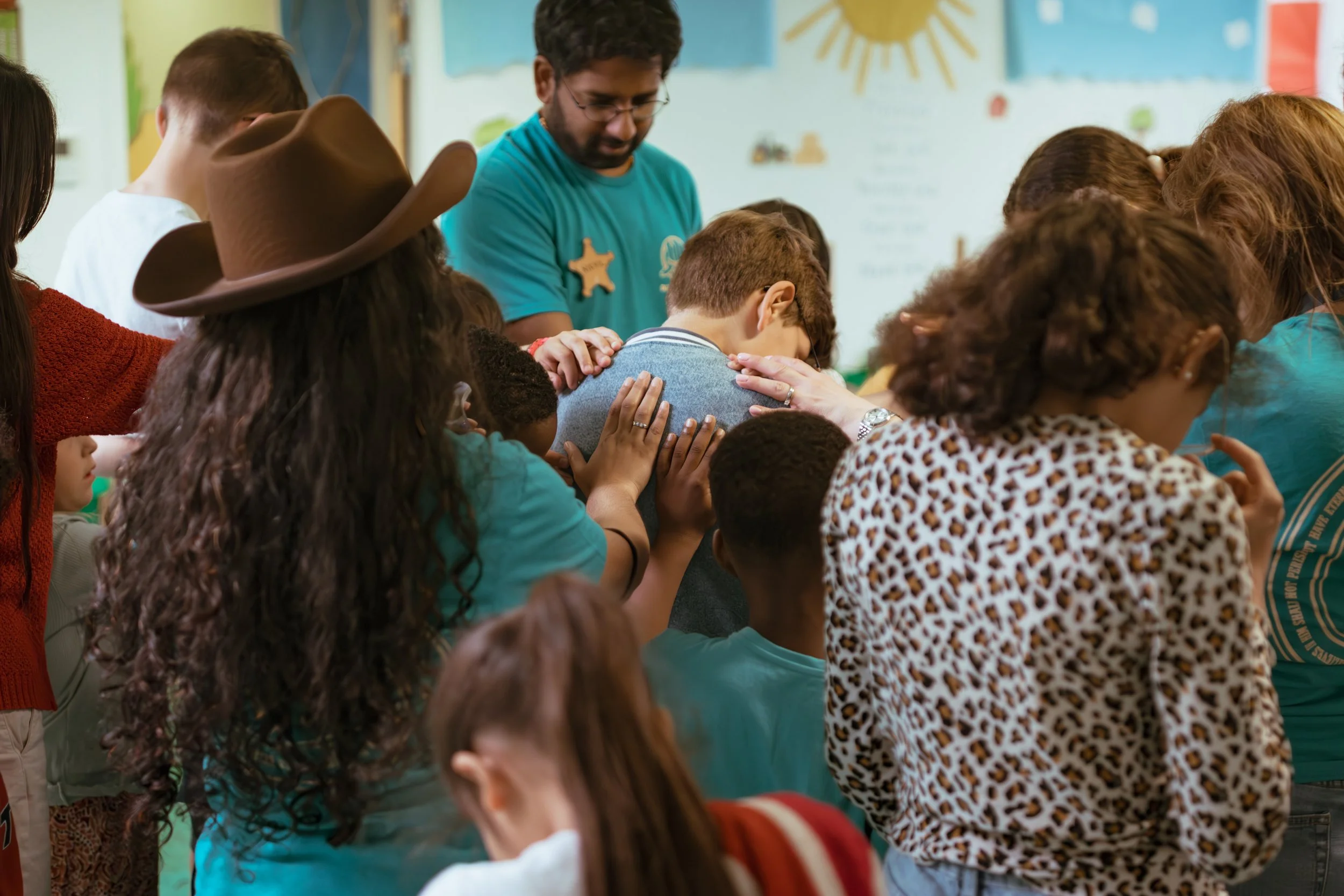 A group of children and an adult in a classroom engaged in prayer or a moment of reflection, with hands on each other's backs and shoulders, fostering a sense of community and spirituality.