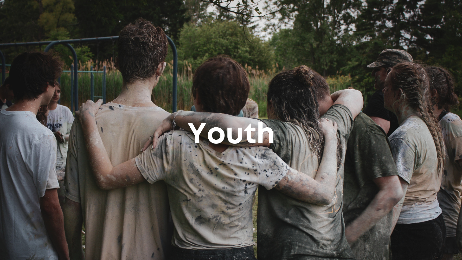 Group of young people with muddy clothes standing together outdoors in a circle with arms around each other, at a park or field with trees in the background, captioned "Youth".