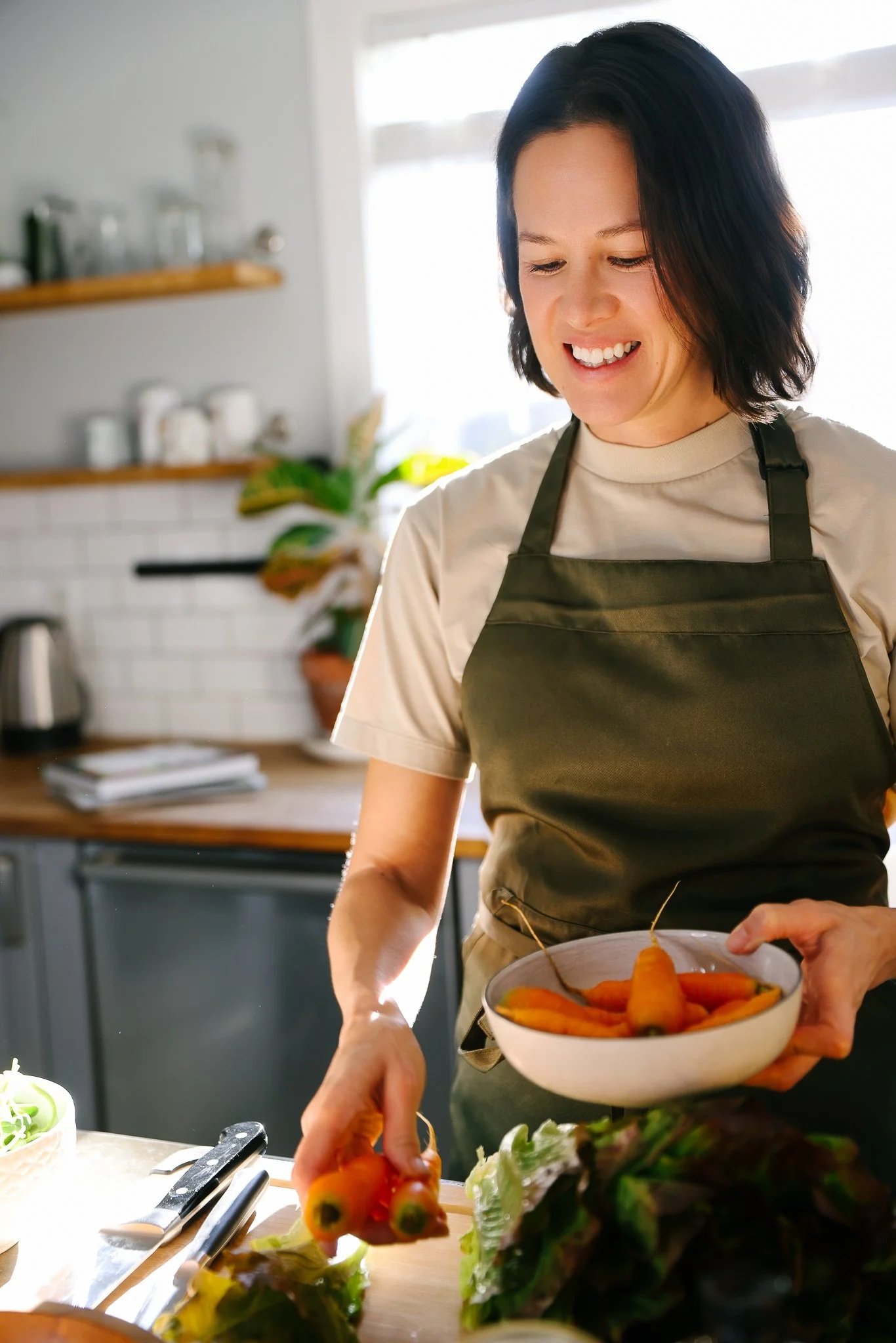 A woman with long dark hair, wearing a green apron and beige top, in the kitchen with fresh veggies