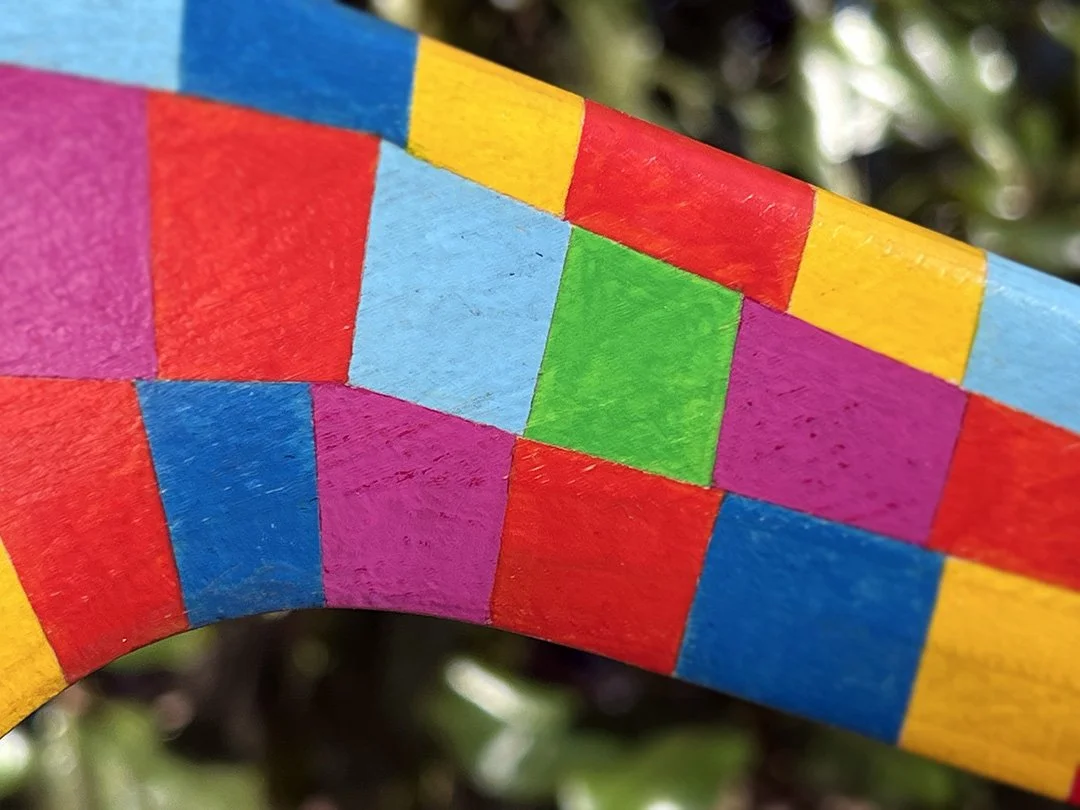 Close-up of a brightly painted wooden boomerang with colorful squares in red, blue, green, yellow, and purple, set against a blurred outdoor background.