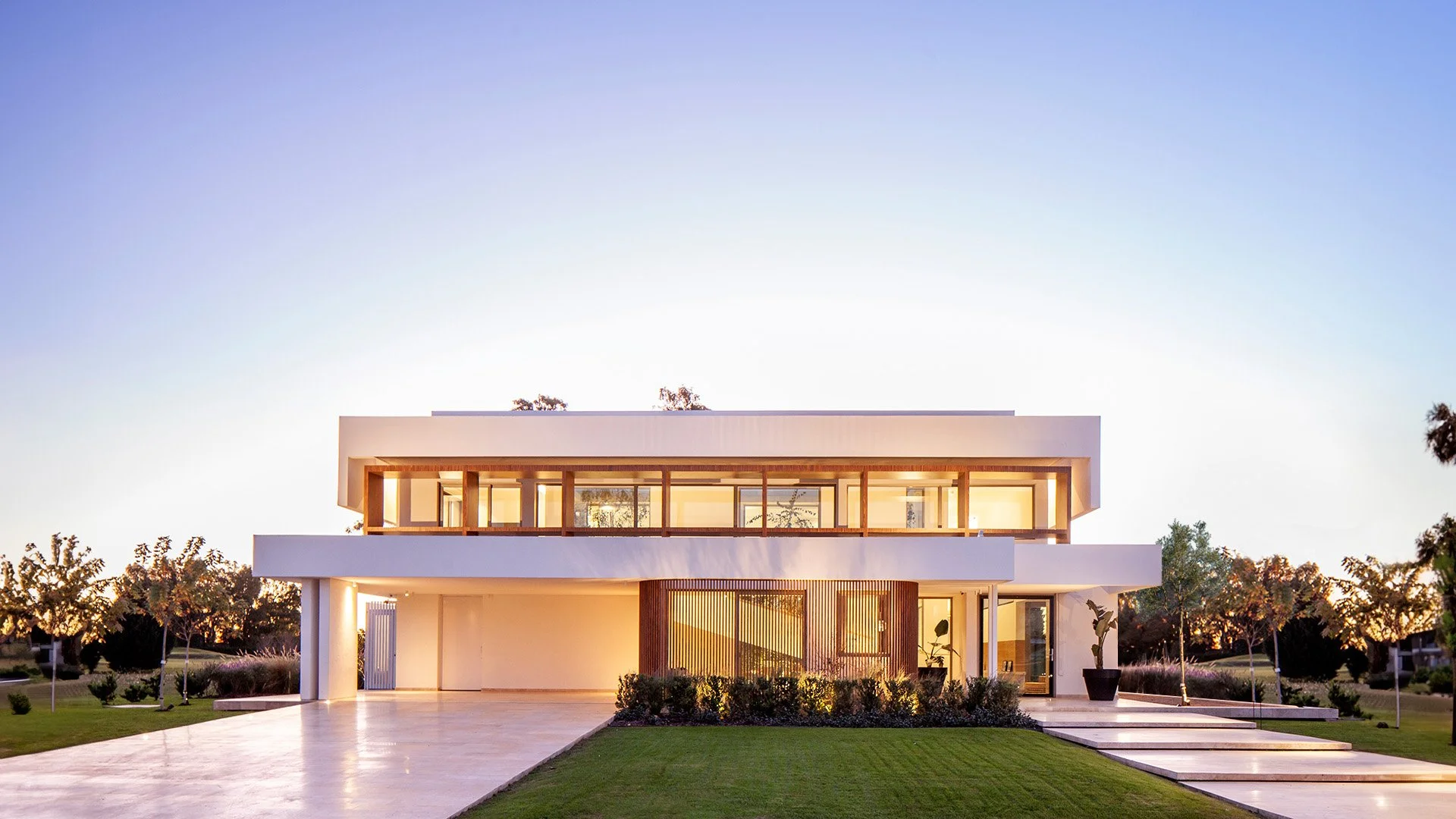Casa moderna de dos pisos con paredes blancas, ventanas grandes, jardín con césped y árboles pequeños, en un atardecer
