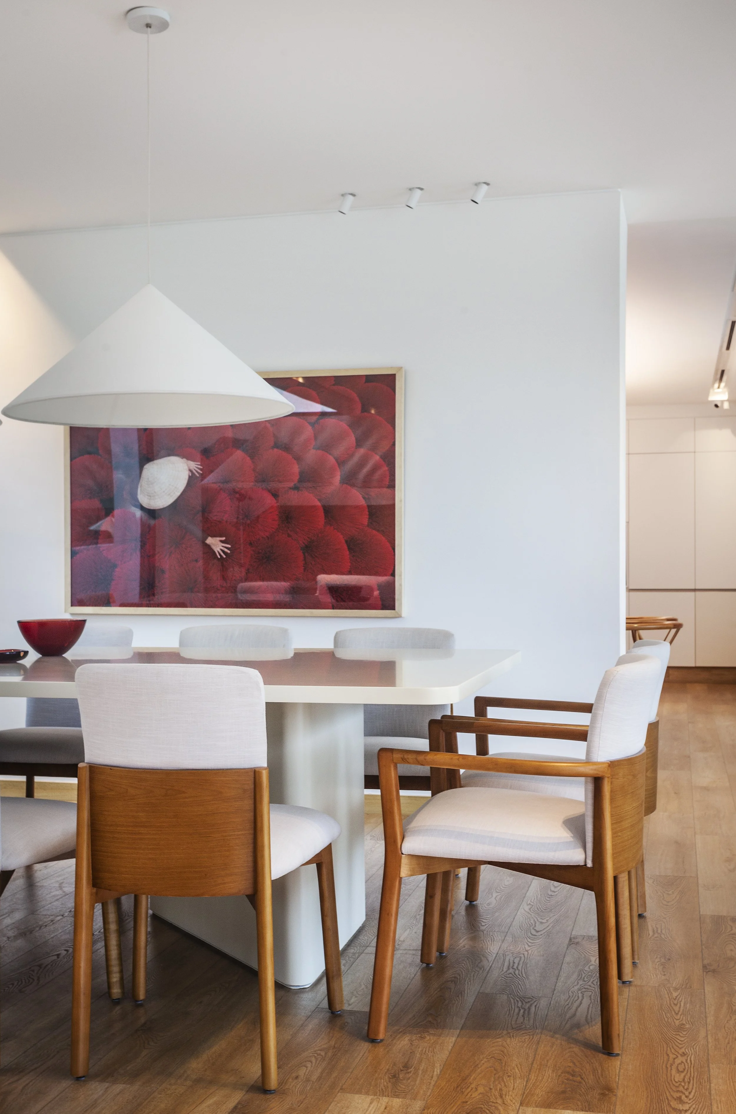 Comedor con mesa blanca rodeada de sillas de madera y tapizado claro, en una pared hay un cuadro con un fondo rojo y una figura de sombrero y manos. Hay una lámpara blanca grande colgando del techo.