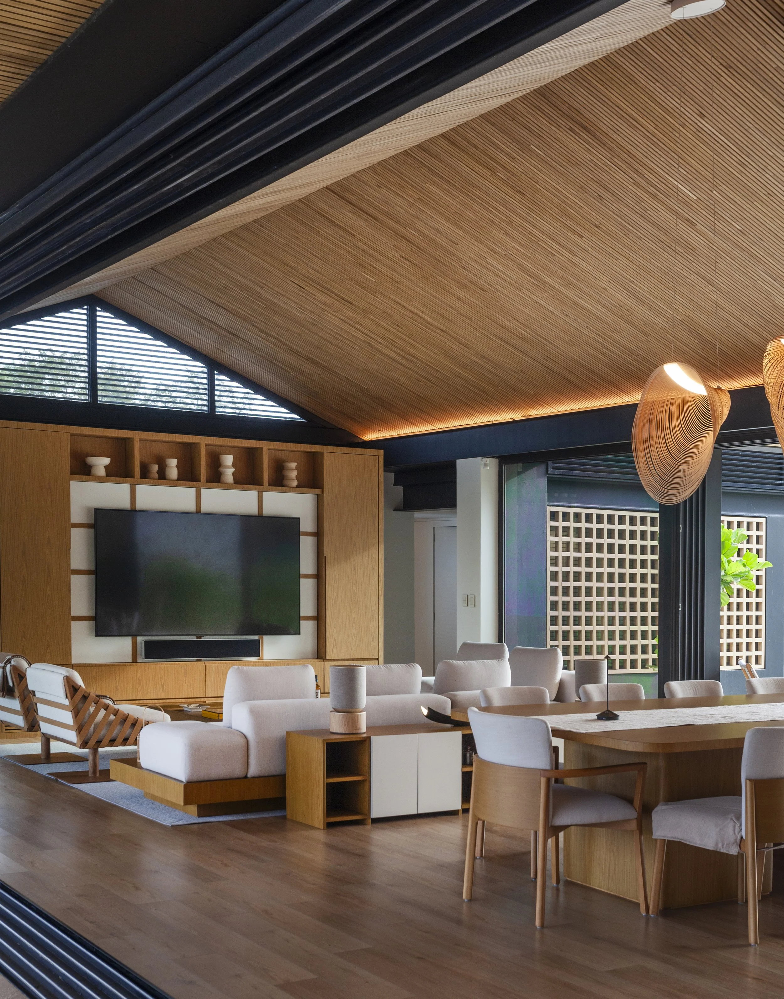 Sala de estar moderna con muebles de madera clara, paredes decoradas con madera y un techo de madera en pendiente. Tiene un televisor grande en la pared, un sofá beige y una mesa de comedor con sillas de madera y cojines claros.