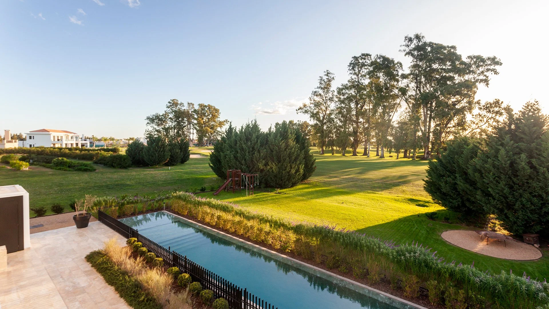 Vista de jardín con área de piscina, árboles grandes, césped, y estructura de juegos infantiles al atardecer.