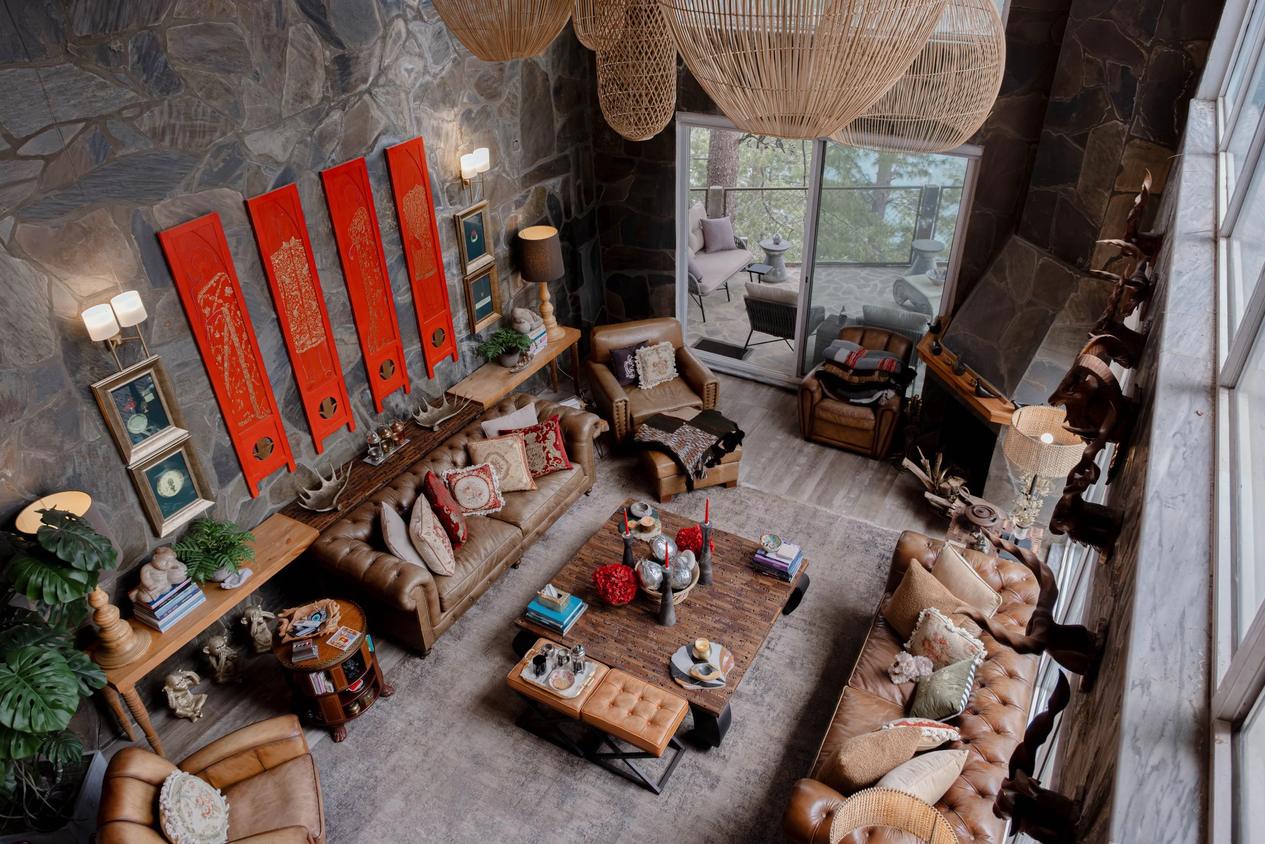 A high-angle view of a cozy living room with a stone wall, leather sofas, colorful pillows, a wooden coffee table with decorative items, framed artwork, plants, and large hanging light fixtures. There's a sliding glass door leading to a balcony with outdoor seating and green trees outside.