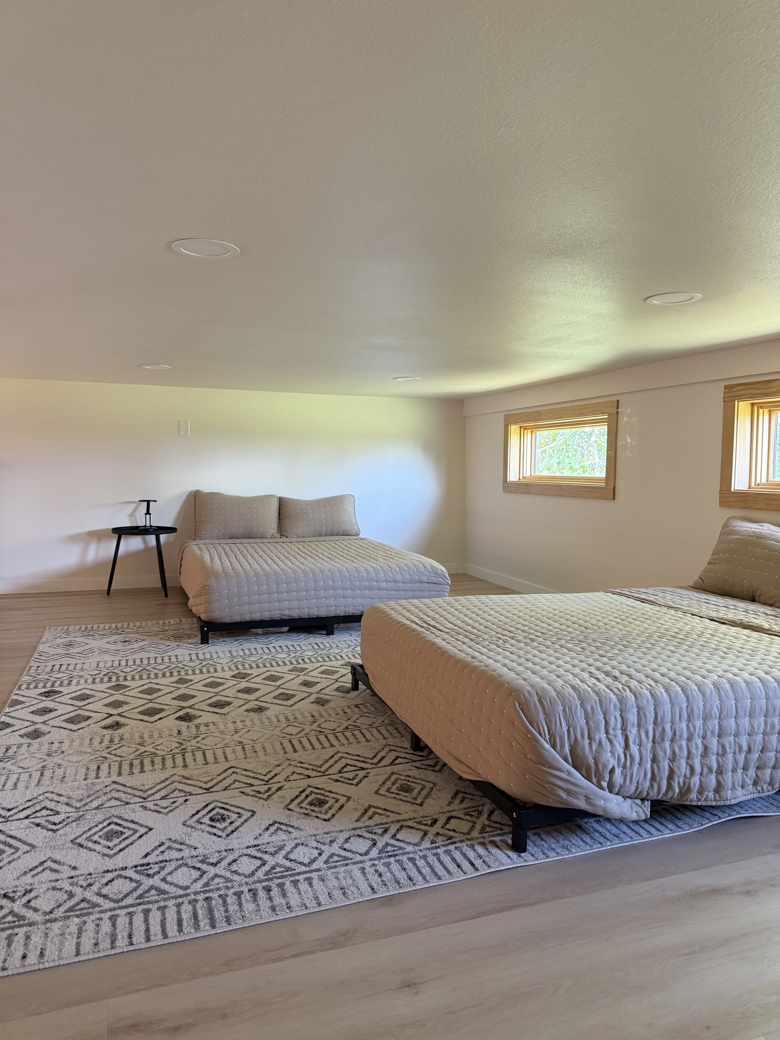 A spacious bedroom with two beds, a small black side table next to one bed, a patterned rug on the wooden floor, and two small windows with wooden frames letting in natural light.