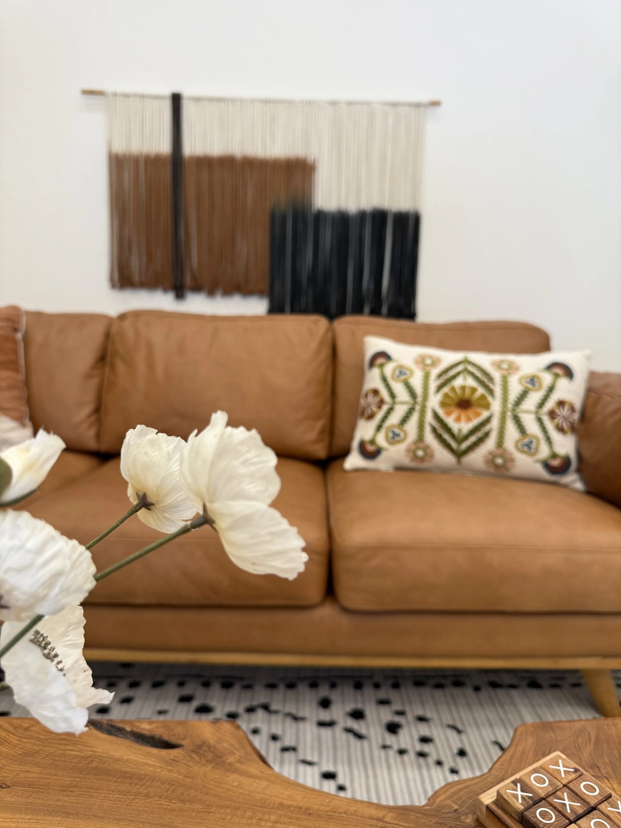 Living room with a tan leather sofa, a decorative pillow with an embroidered design, abstract wall art made of vertical strings in neutral, brown, and black colors, a patterned rug, a wooden coffee table with a tic-tac-toe game, and white flowers in the foreground.
