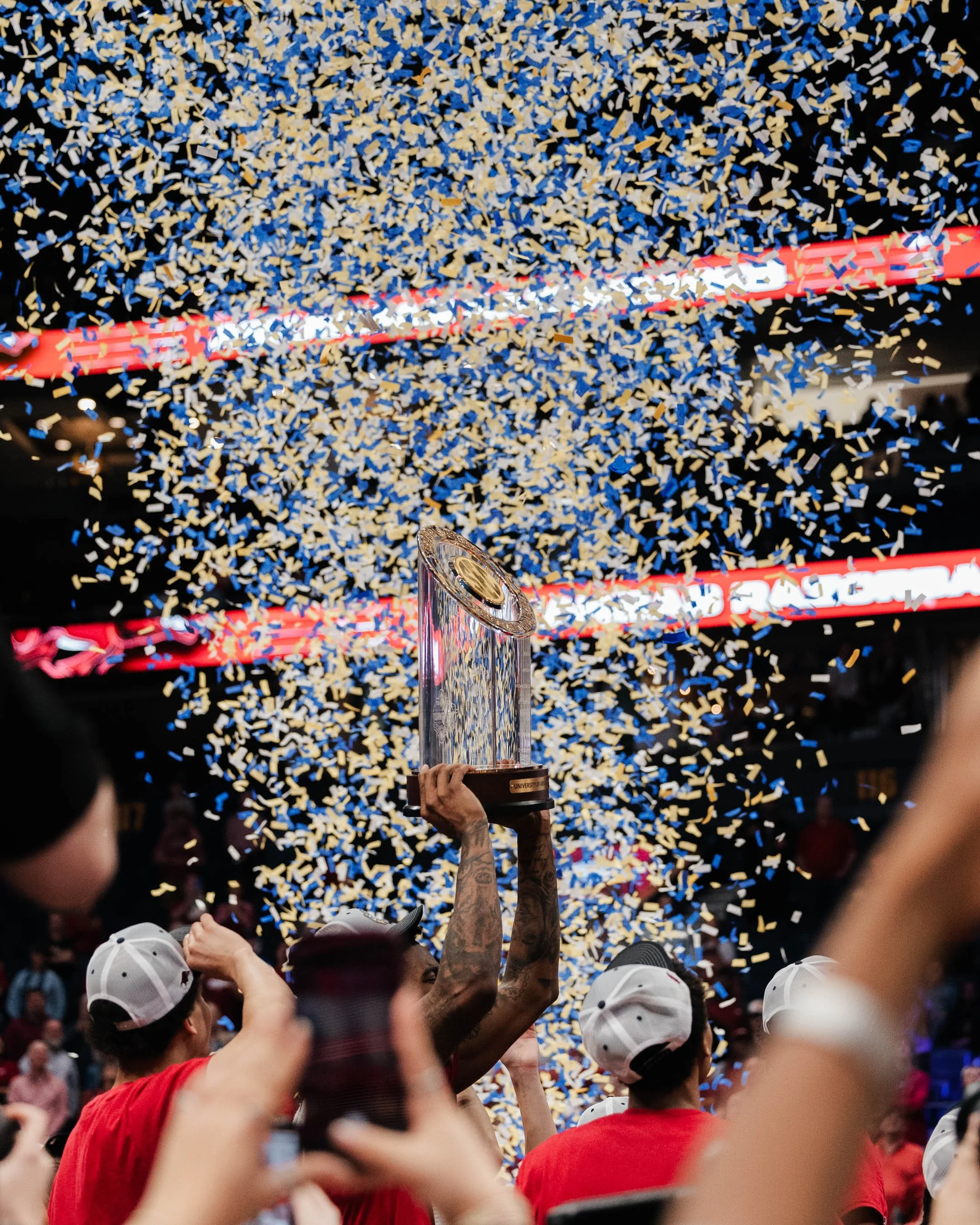 People celebrating with a trophy amidst blue and yellow confetti, wearing red and white caps and shirts, with one person holding the trophy high.