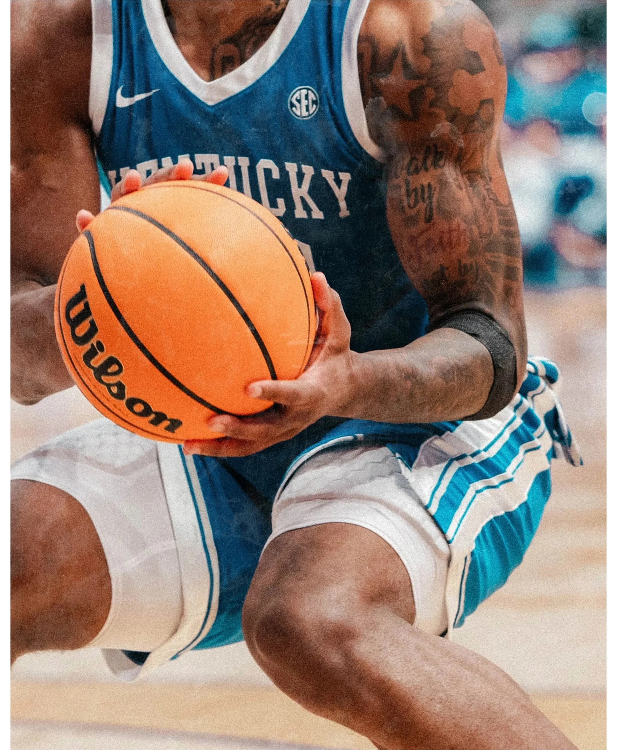 A person in a blue basketball jersey holding an orange basketball. The person has tattoos on both arms and a tattoo on the chest visible through the jersey. The background is blurred with bright lights.