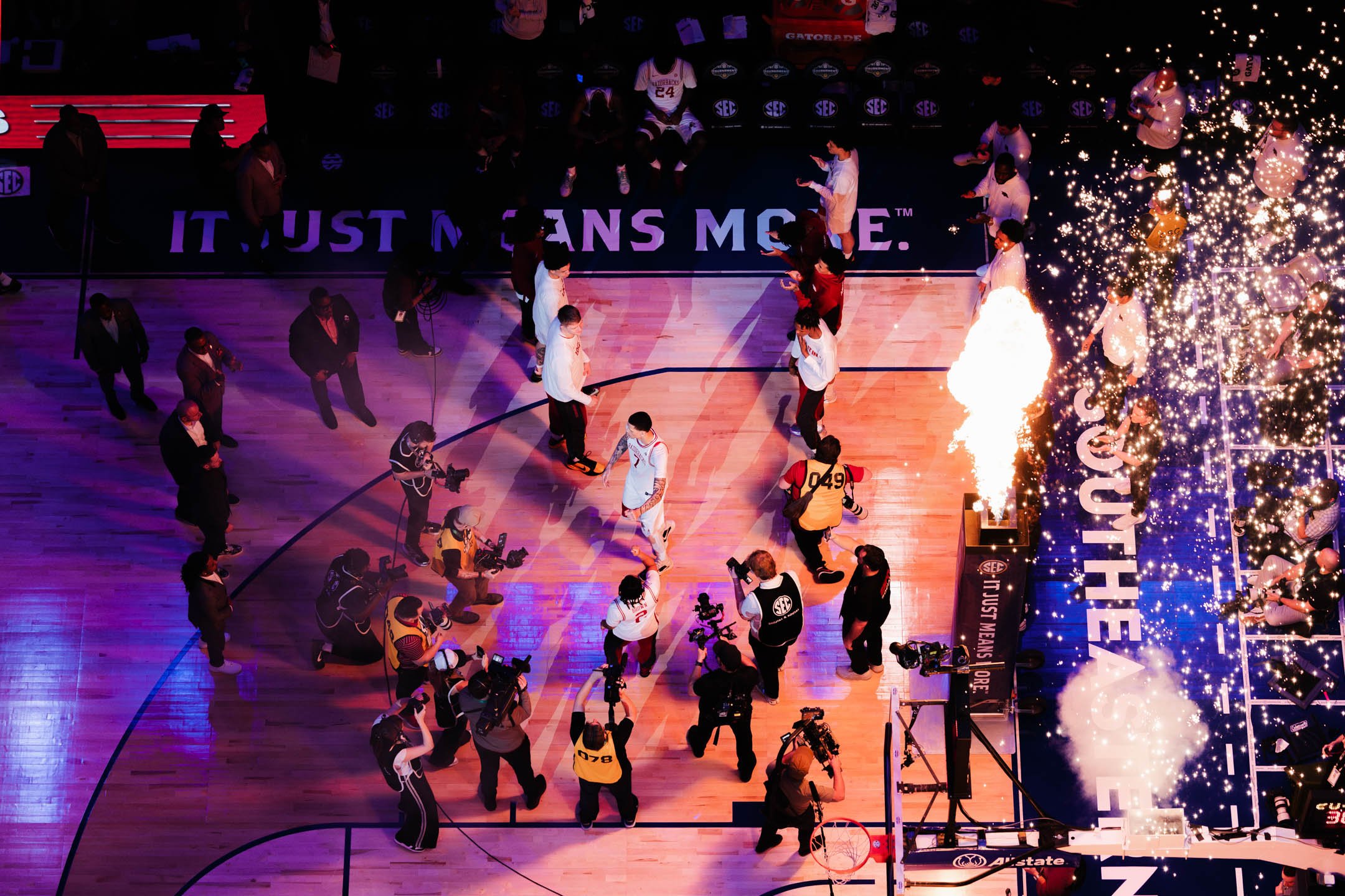An indoor basketball court filled with players, media, and photographers, with a Christmas tree decorated with lights and ornaments on the right side. The court has the NBA SEC sponsorship banners and a large advertisement board.