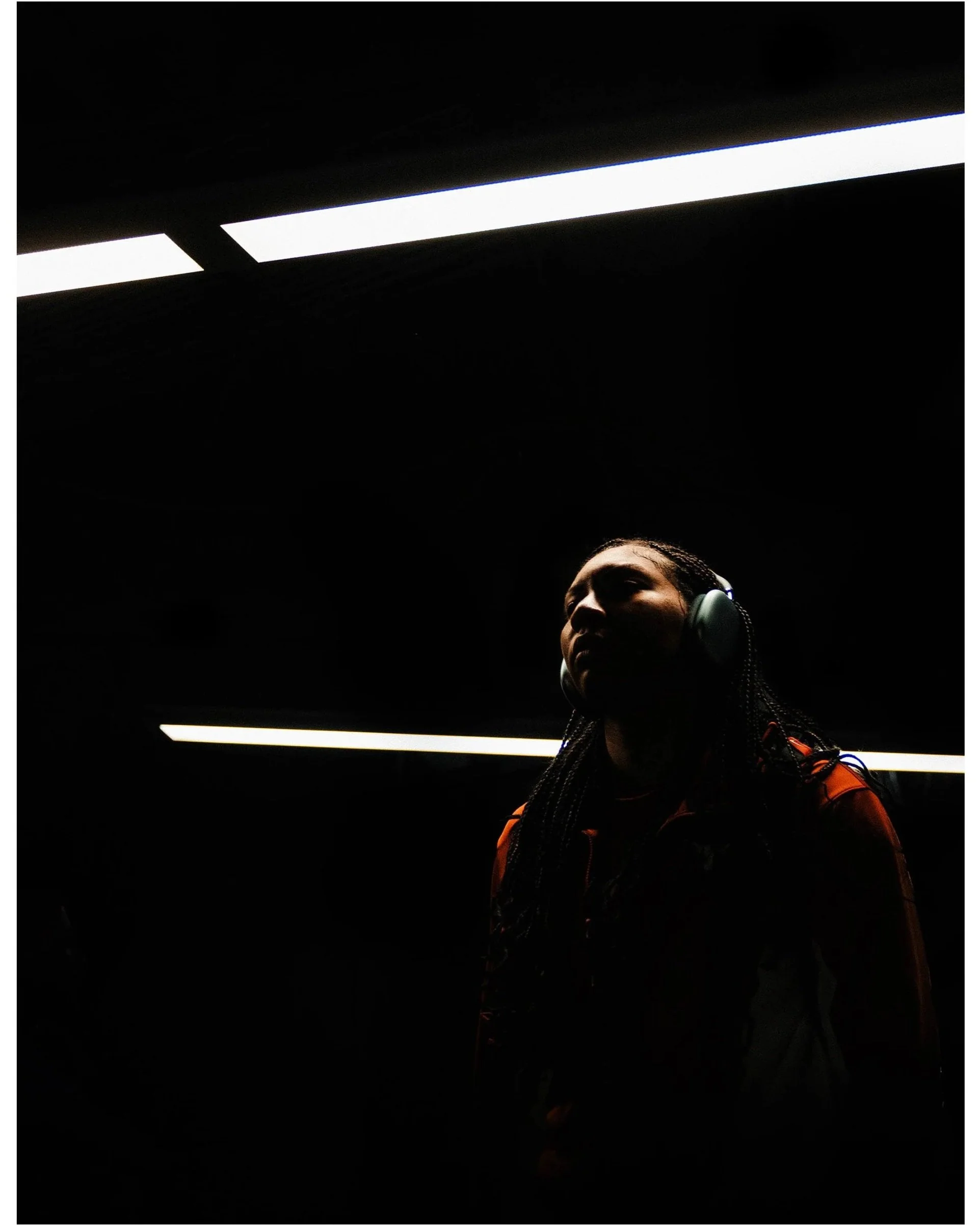 A woman with dark braided hair, wearing headphones and an orange jacket, standing in front of a dark background with horizontal fluorescent lights.