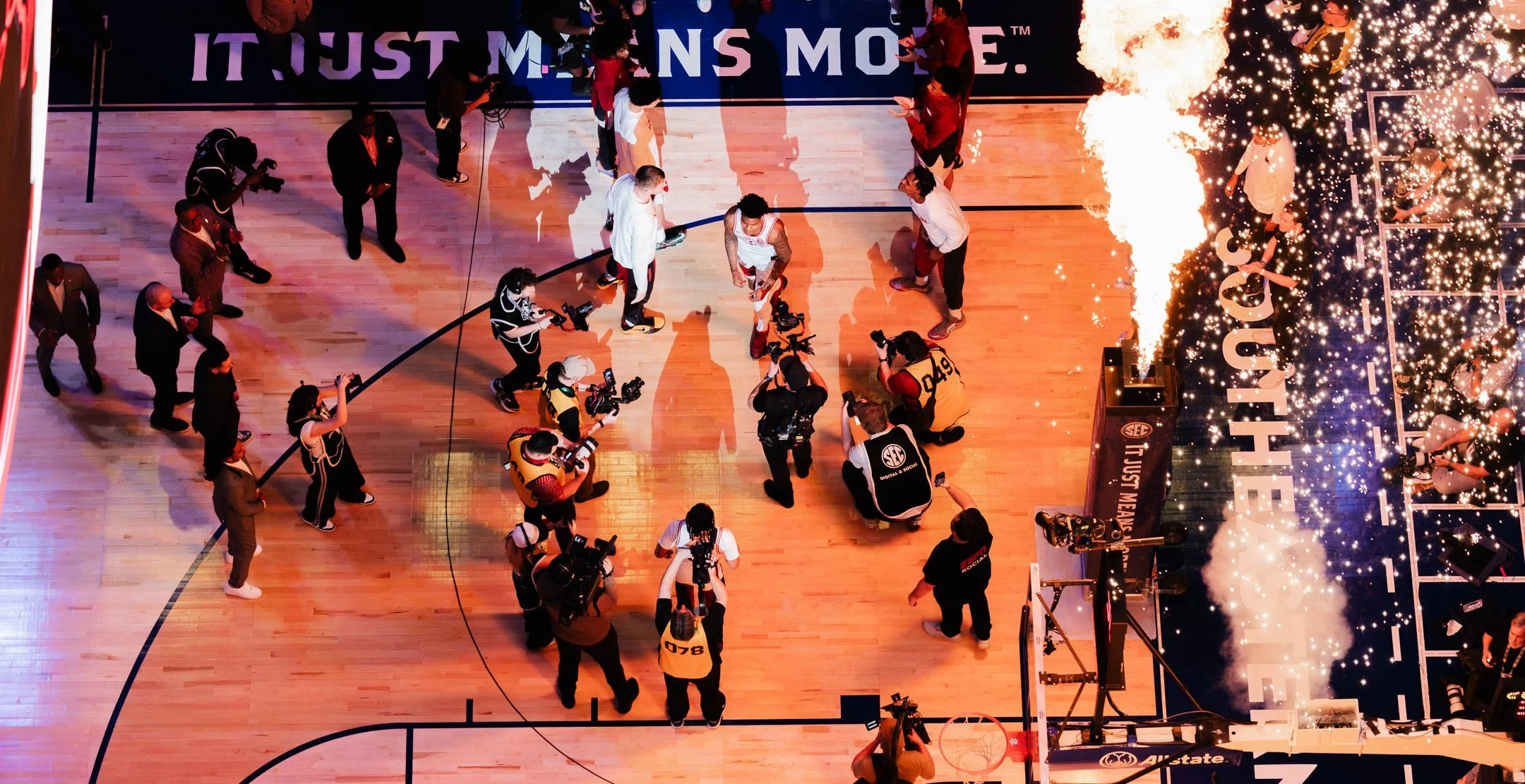 A celebration in a gymnasium with people, including athletes and media, around a basketball court. Fireworks and confetti are present, and a banner reads 'IT JUST MEANS MORE'.