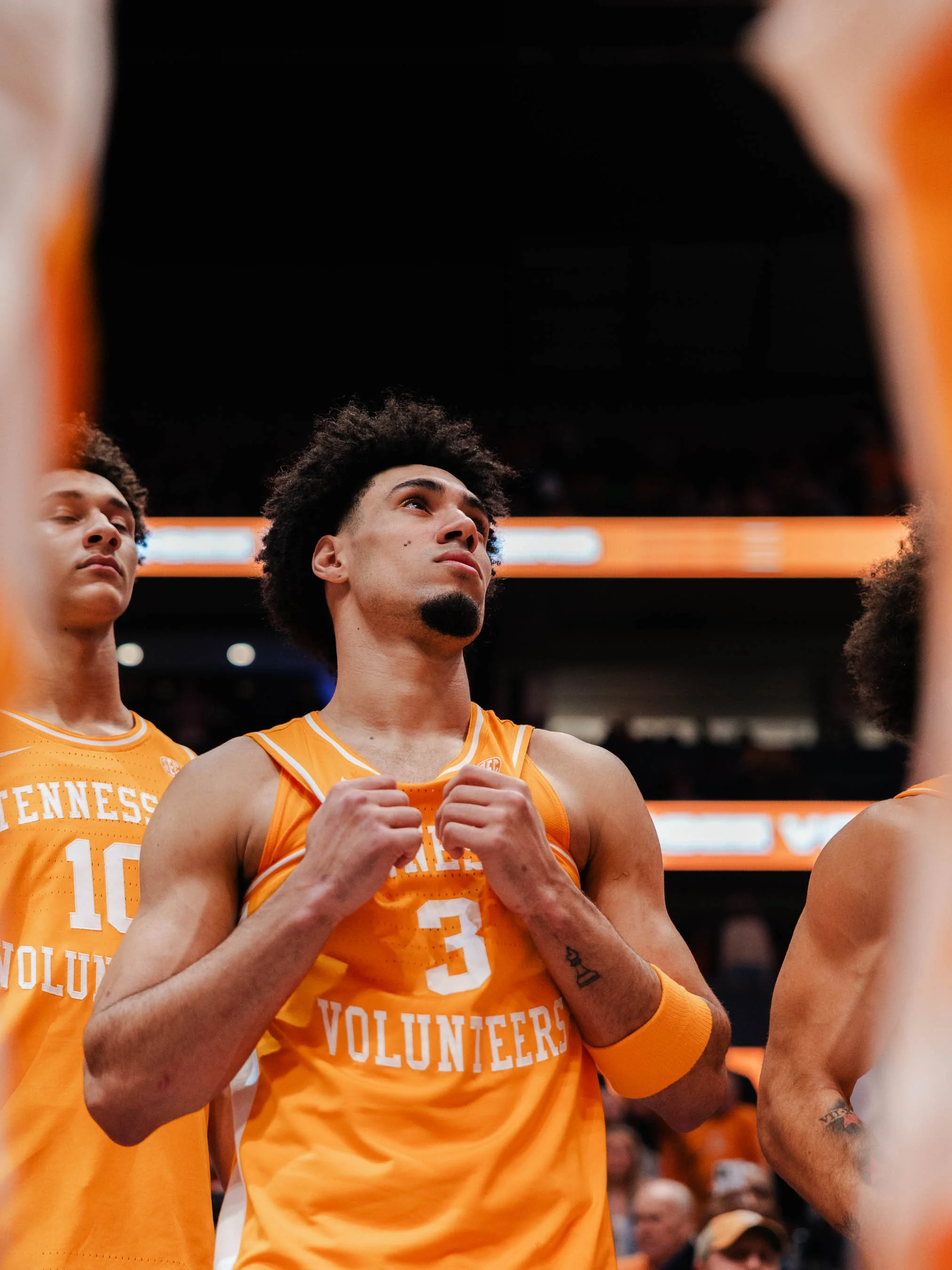 Young male basketball player with curly hair and a goatee, wearing an orange volunteering jersey, standing on the court with hands clasped, surrounded by teammates during a team moment.