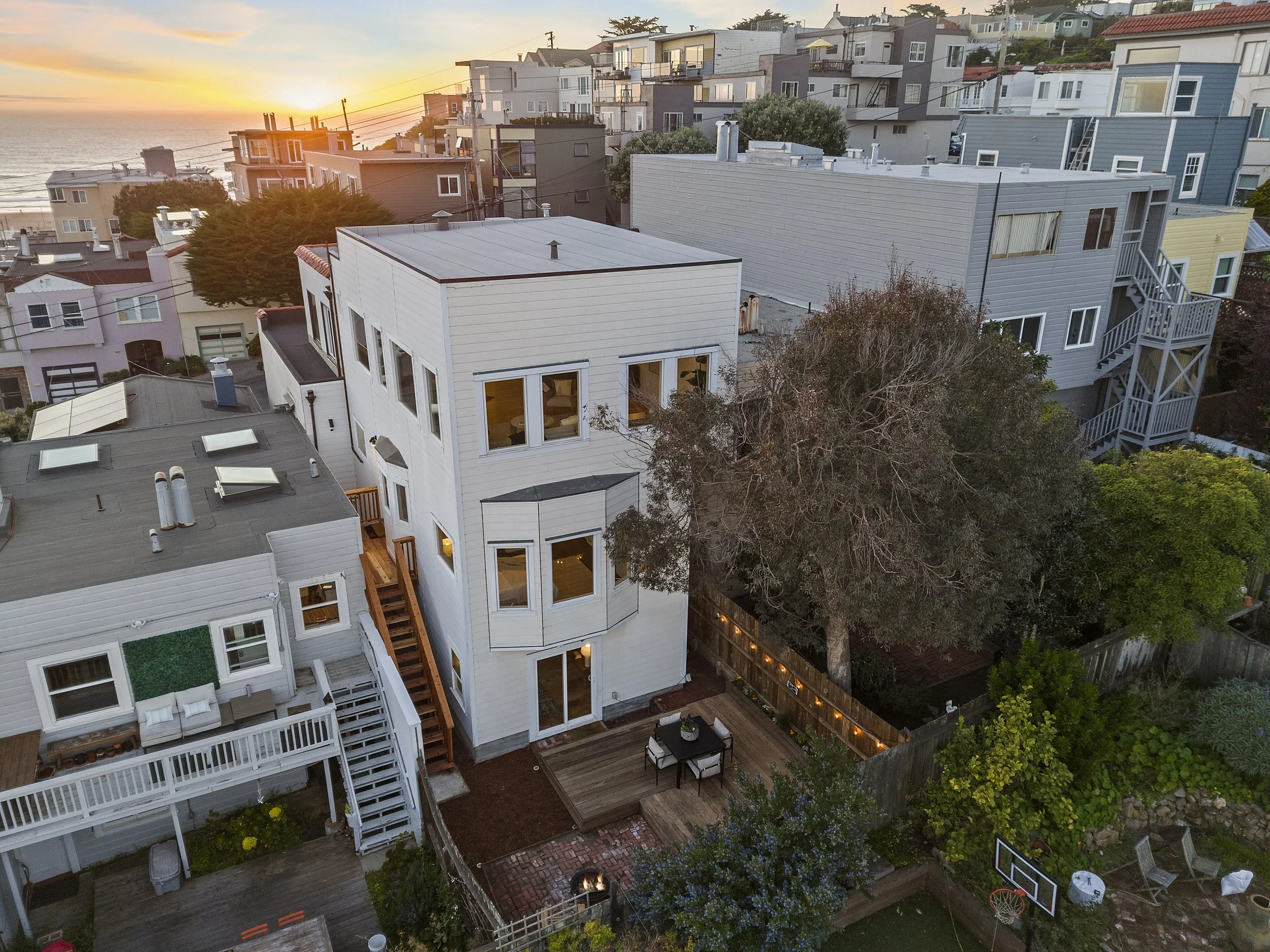 Aerial view of a residential neighborhood at sunset, featuring modern multi-story houses with decks, trees, and a backyard with a fire pit and outdoor furniture.