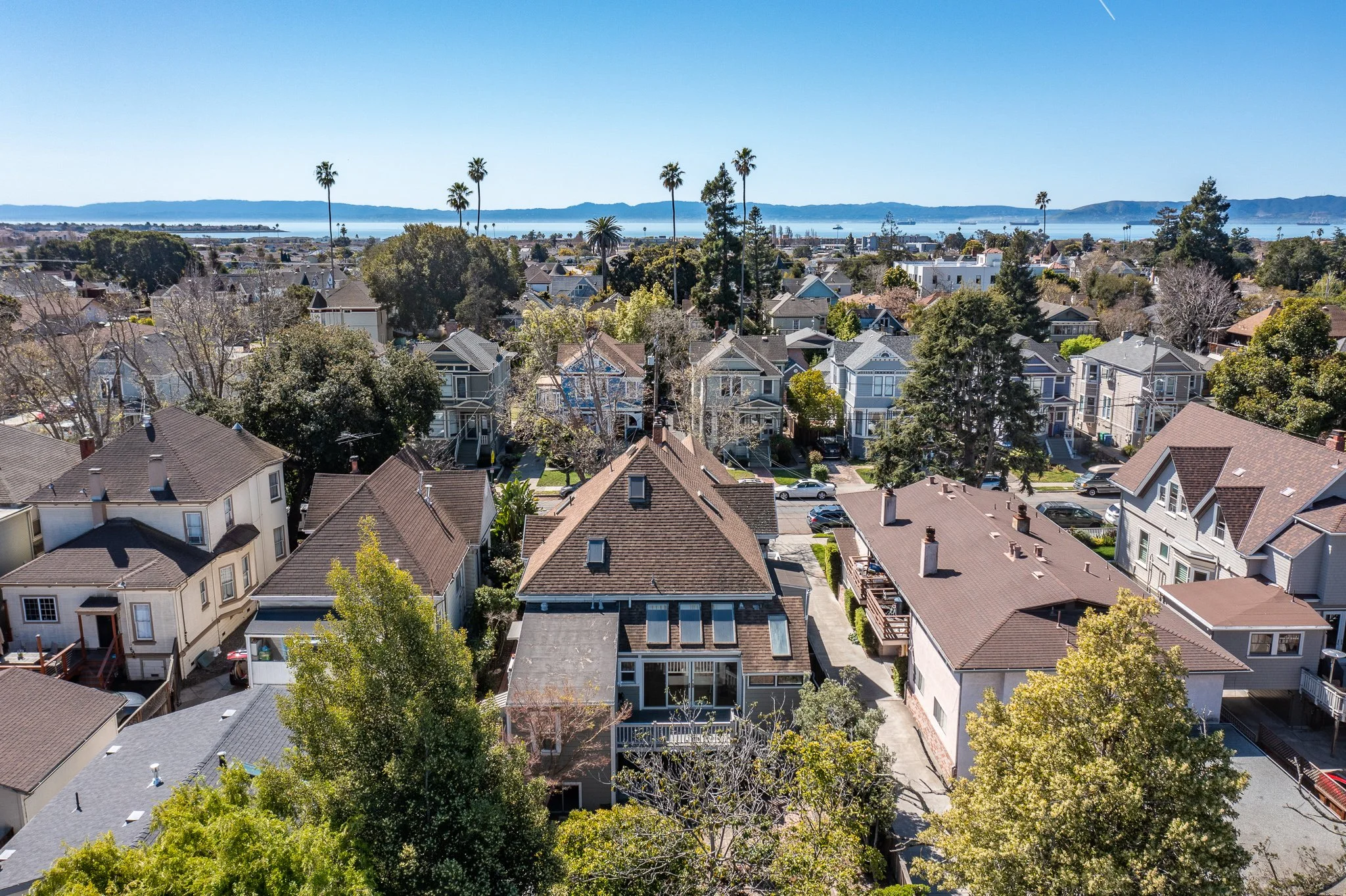 Aerial view of a residential neighborhood with houses, trees, and streets, overlooking a body of water with mountains in the distance under a clear blue sky.