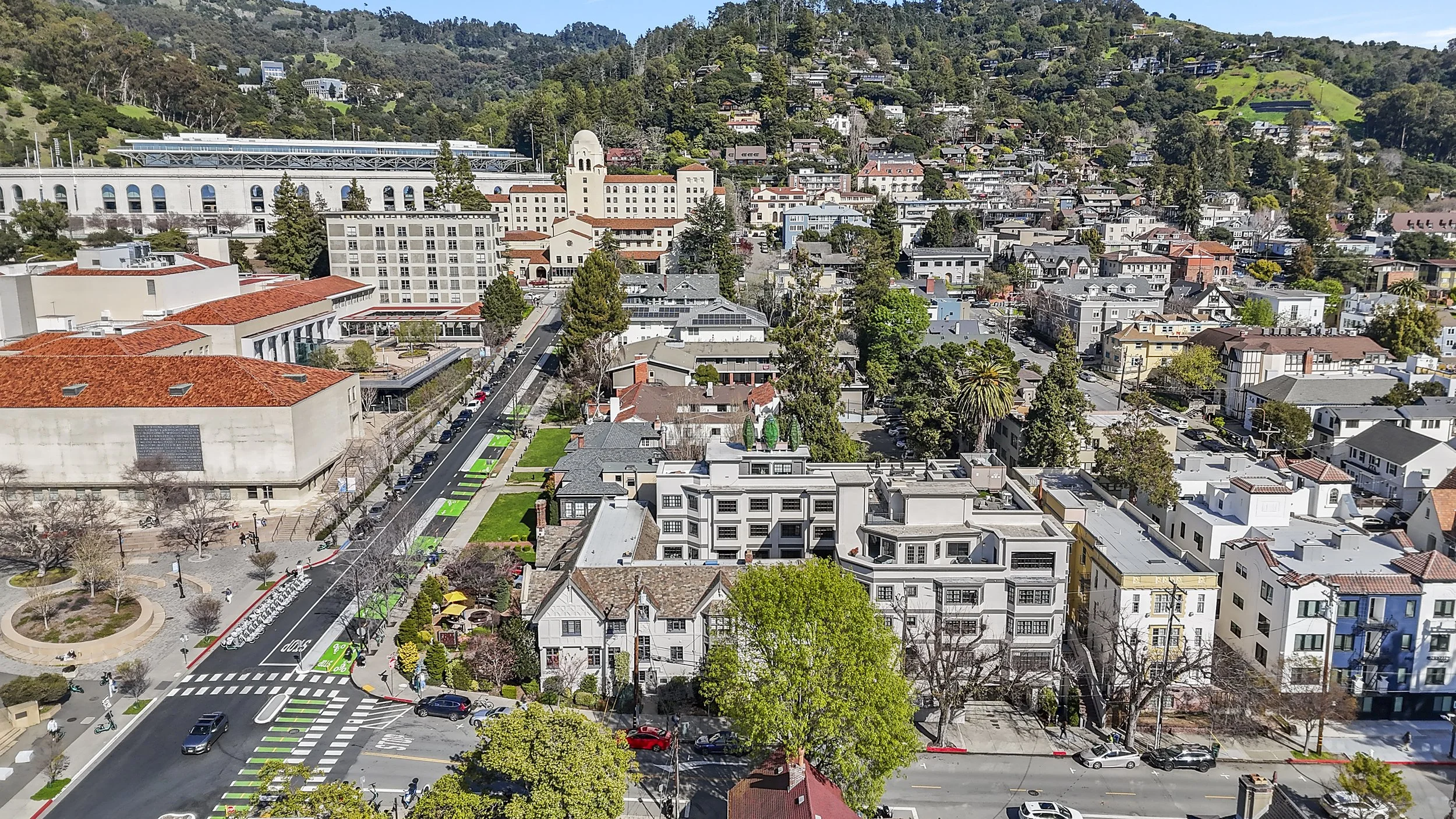 Aerial view of a city with a mix of modern and historic buildings, green hills in the background, streets, trees, and a park with a circular walkway.