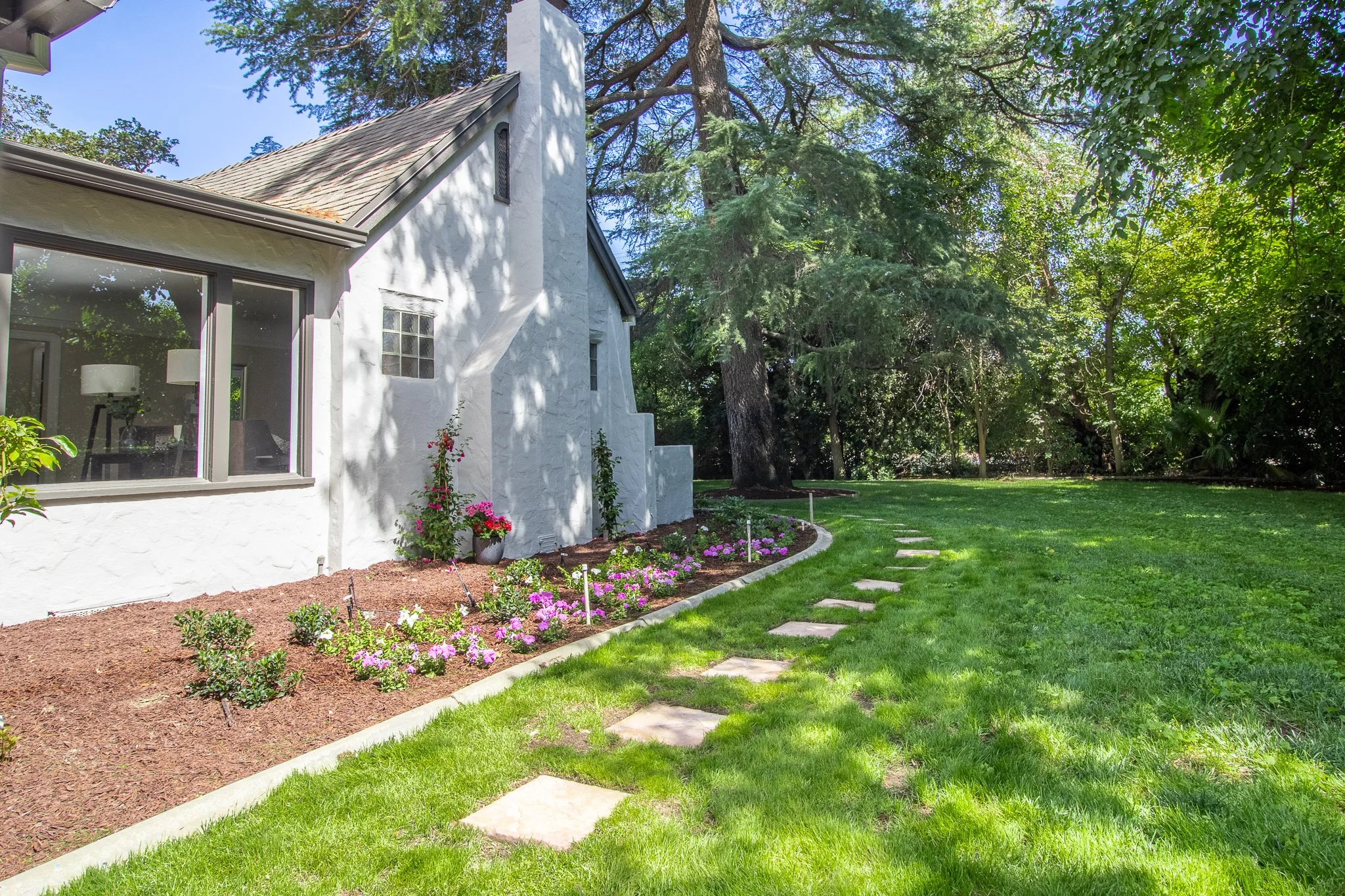 View of a backyard with a white house and large windows on the left, a well-maintained lawn with a stone pathway on the right, flower bed with pink and purple flowers near the house, and tall trees providing shade in the background.